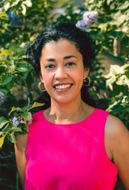 A woman in a pink tank top is smiling in front of a bush.