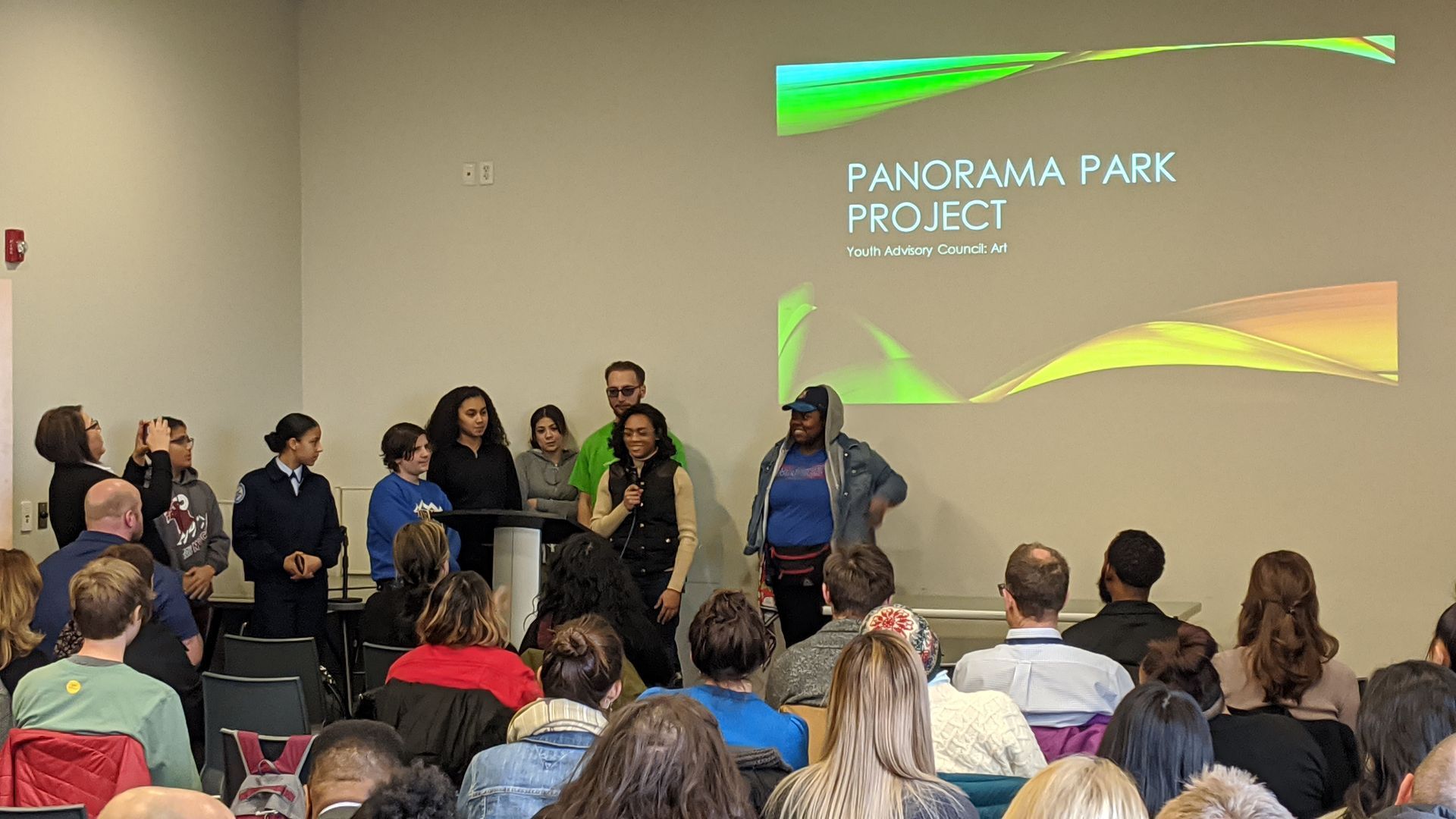 Youth speak to a large group of people with a powerpoint in the background