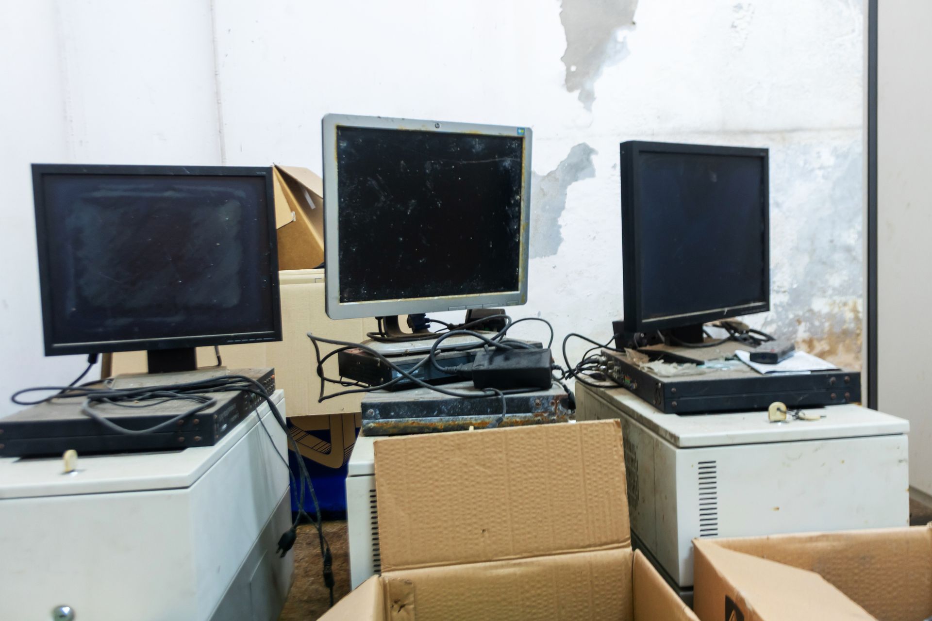 Three dusty computer monitors on white cabinets, with wires and boxes in a cluttered room.