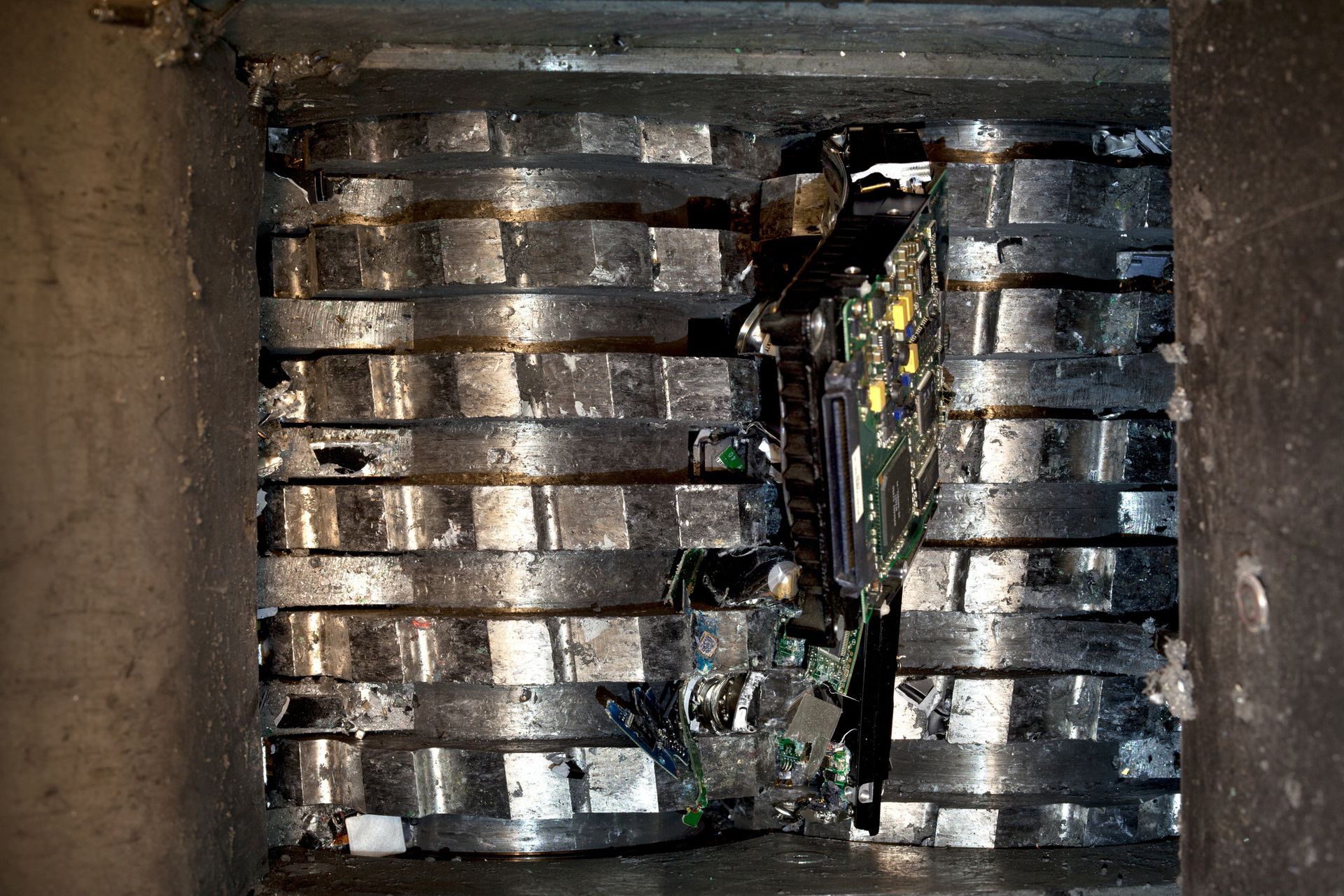 A circuit board being shredded by industrial blades inside a metal container.