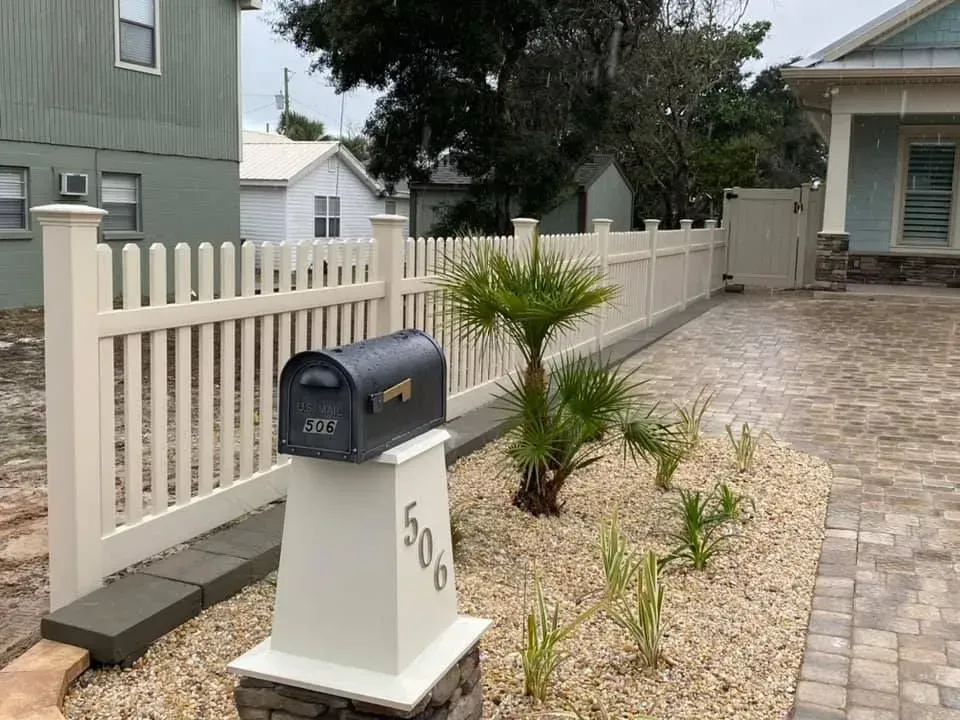 Mailbox on a white post beside a gravel driveway and small plants, next to a white picket fence.