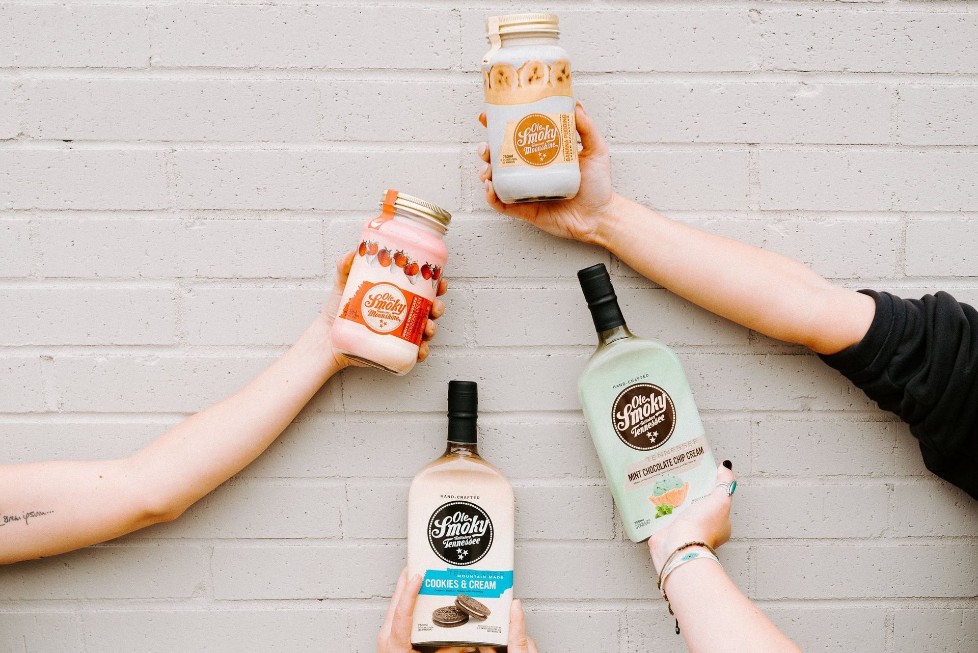 Hands holding up bottles against a white brick wall. Two hands hold jars with drinks, two hands hold liquor bottles.