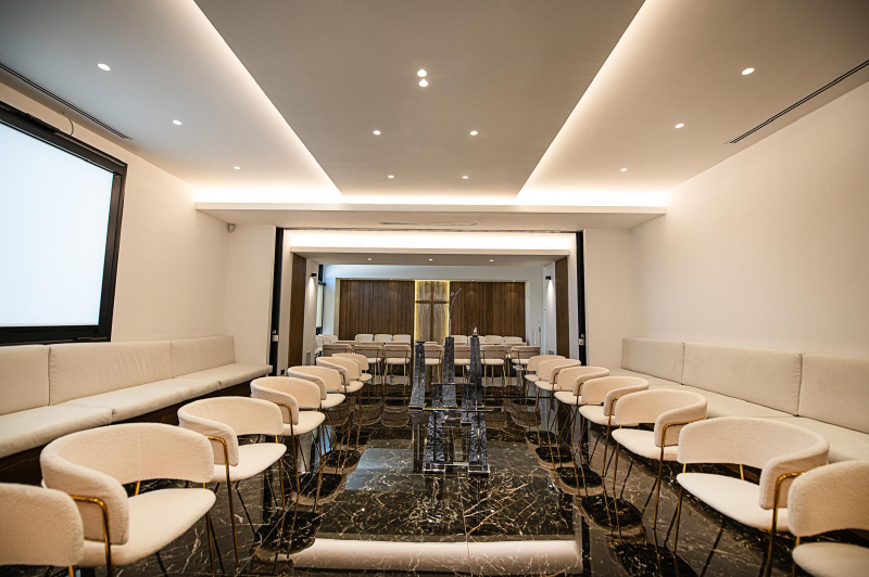 Sala conferenze moderna con sedie bianche, divani beige e pavimento nero lucido illuminato da faretti a incasso.