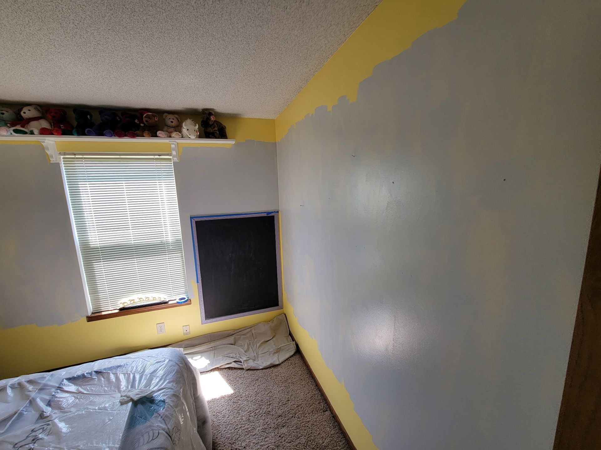 Room with walls partially painted gray, with yellow trim. Bed covered in plastic, window on the left.