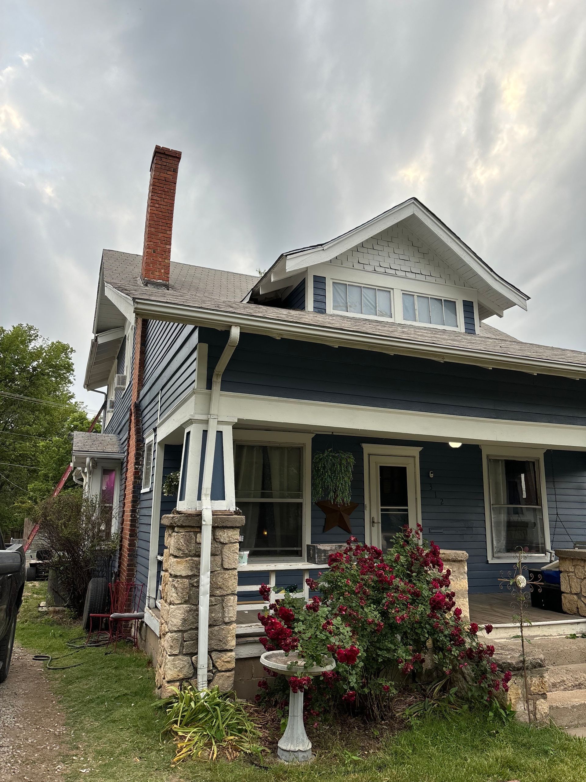 Two-story blue house with white trim, chimney, and cloudy sky. Red rose bush in front.