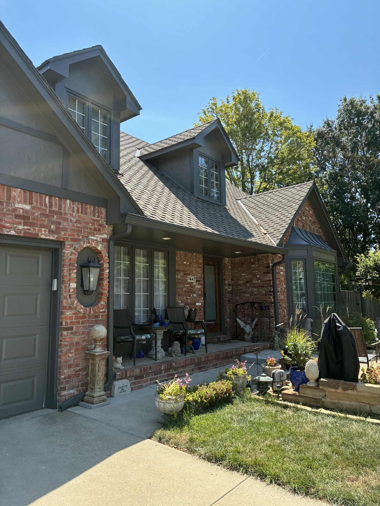 Brick house with gray roof, dormers, and garage.  A front porch with chairs, flower beds, and lawn.