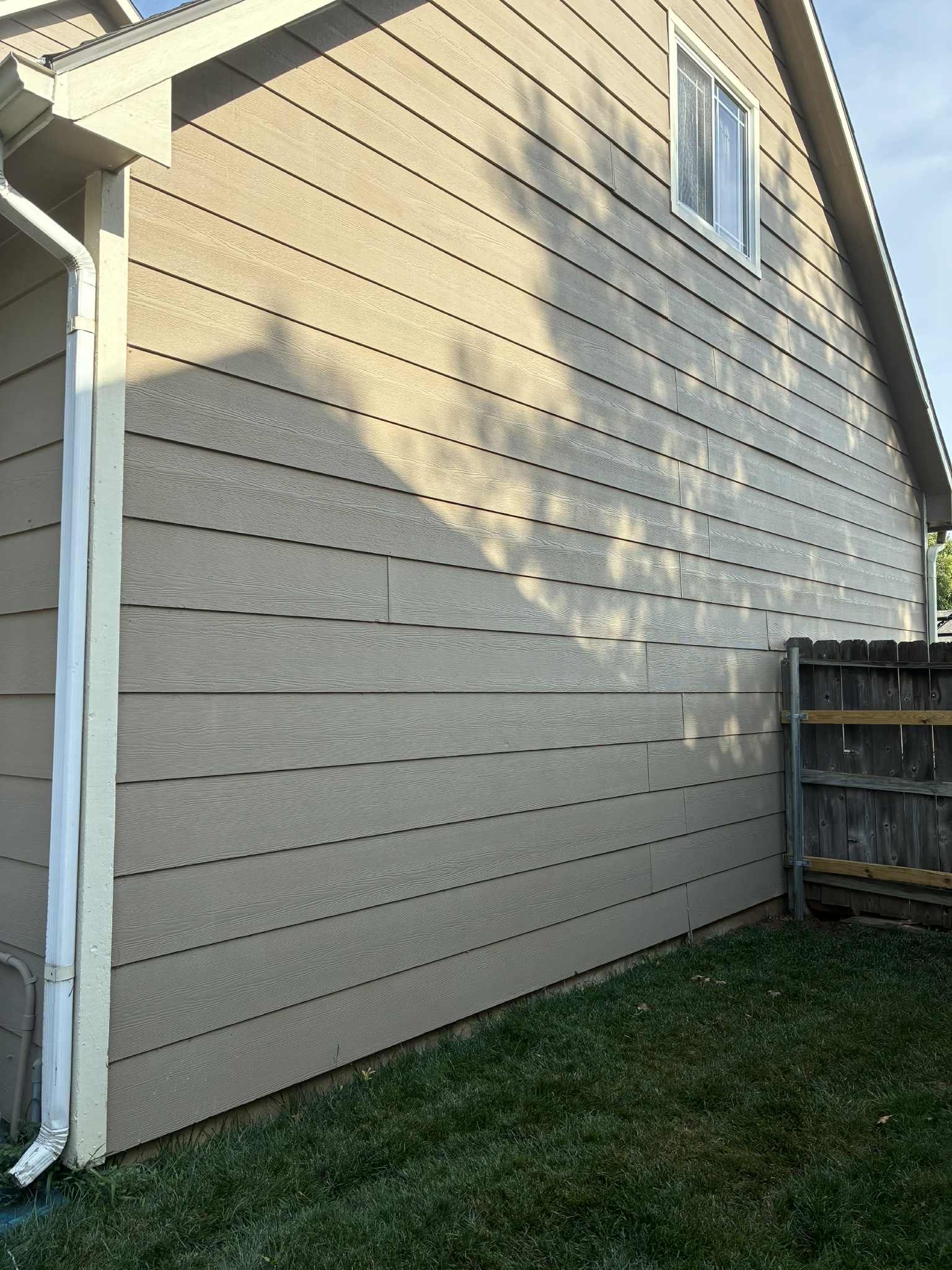 Beige siding of a house with a white window, gutter, and a wooden fence in the yard.