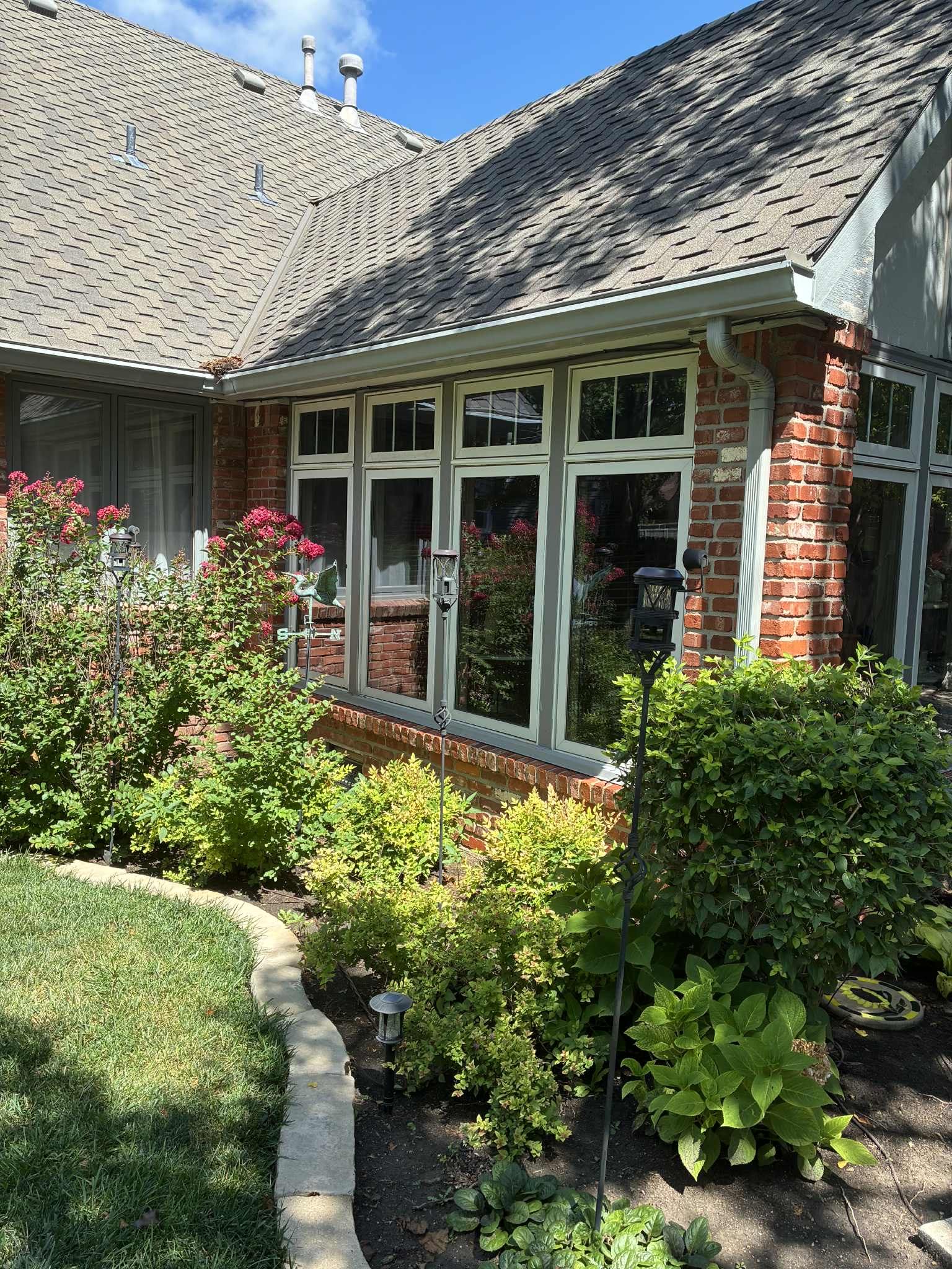 Brick house with a window surrounded by green plants and a curved lawn.