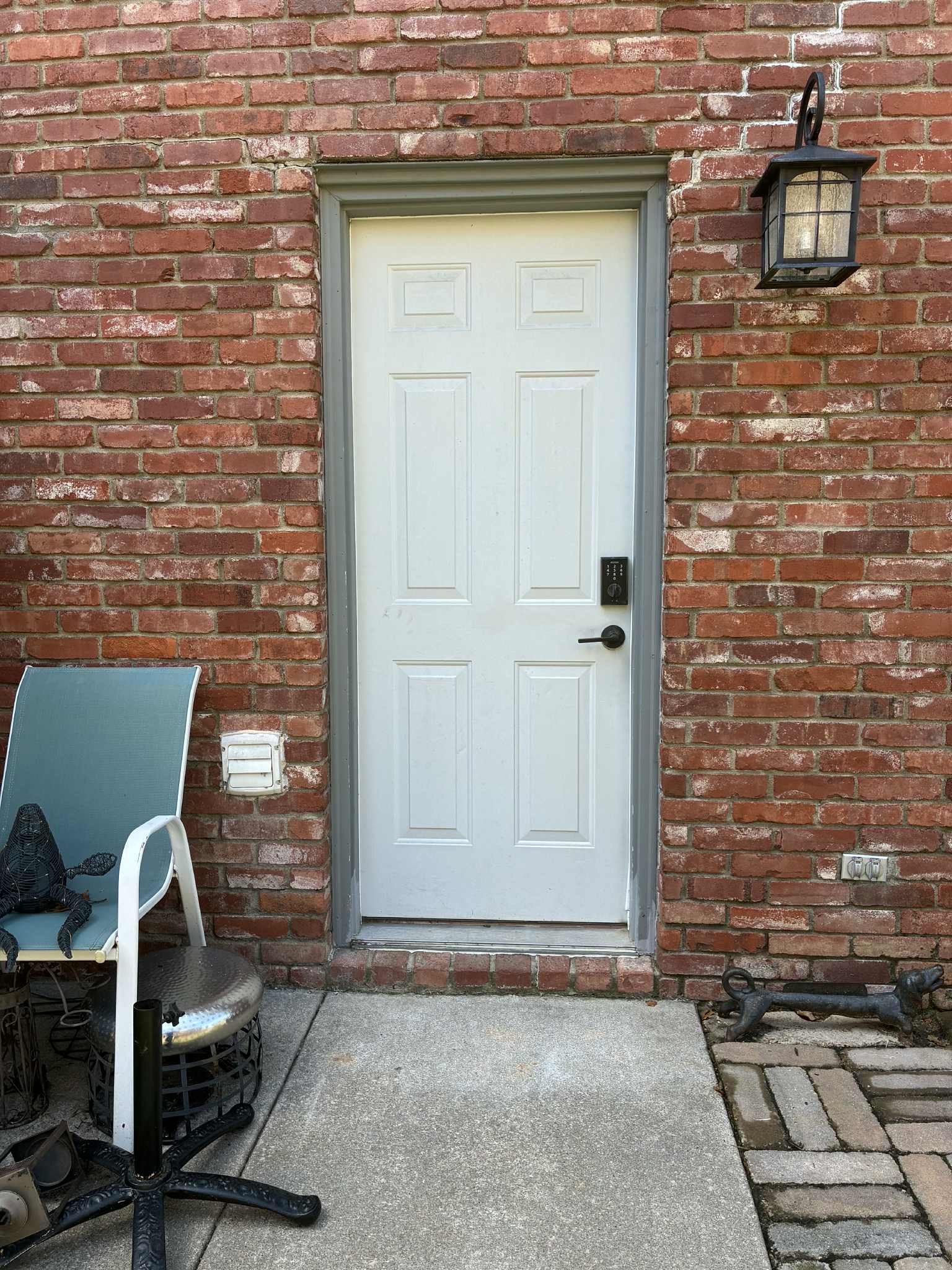 White door set in a brick wall, with a chair to the left and an outdoor light fixture to the right.