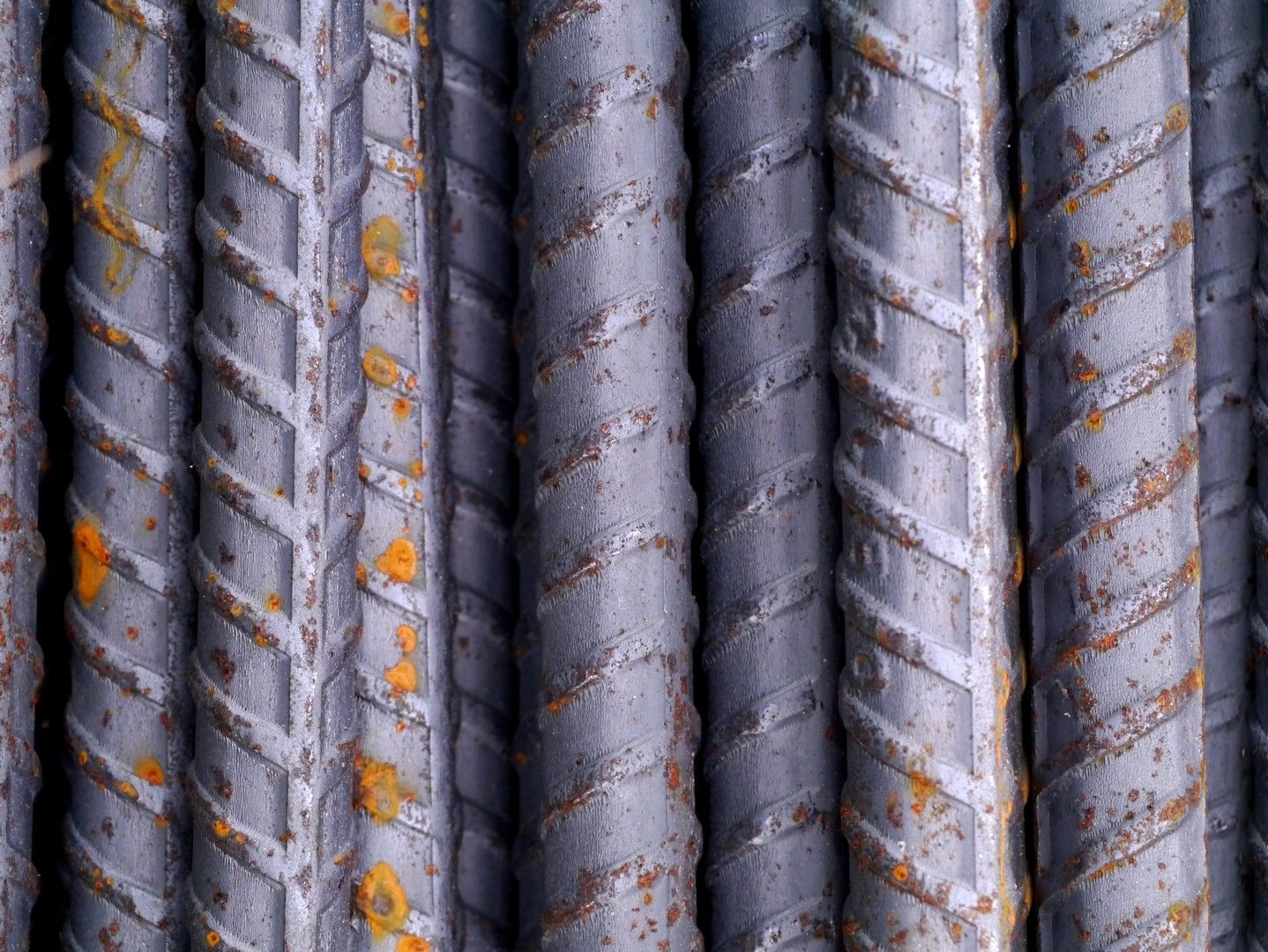 Vista ravvicinata di numerose barre di rinforzo in acciaio nervato di colore grigio scuro, raggruppate insieme e con piccole macchie di ruggine arancione.