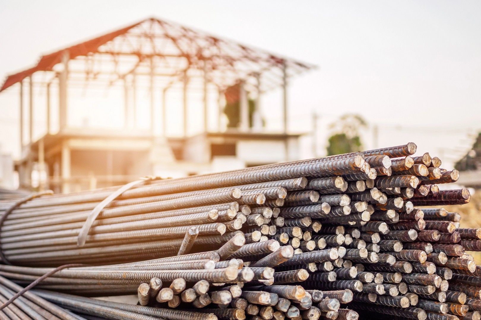 In primo piano, una grande pila di barre d'acciaio per cemento armato con superficie ruvida, mentre sullo sfondo si vede la struttura di un edificio in costruzione ancora incompiuto.