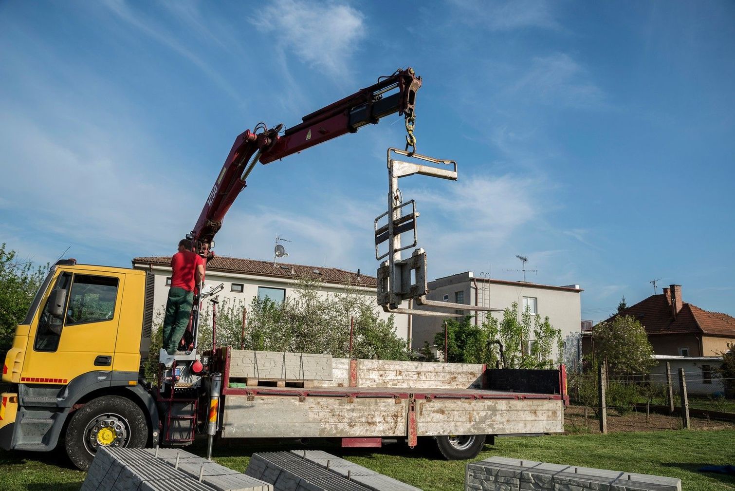 Un camion giallo con gru solleva un carrello elevatore per la posa di mattoni all'aperto, sotto un cielo azzurro.