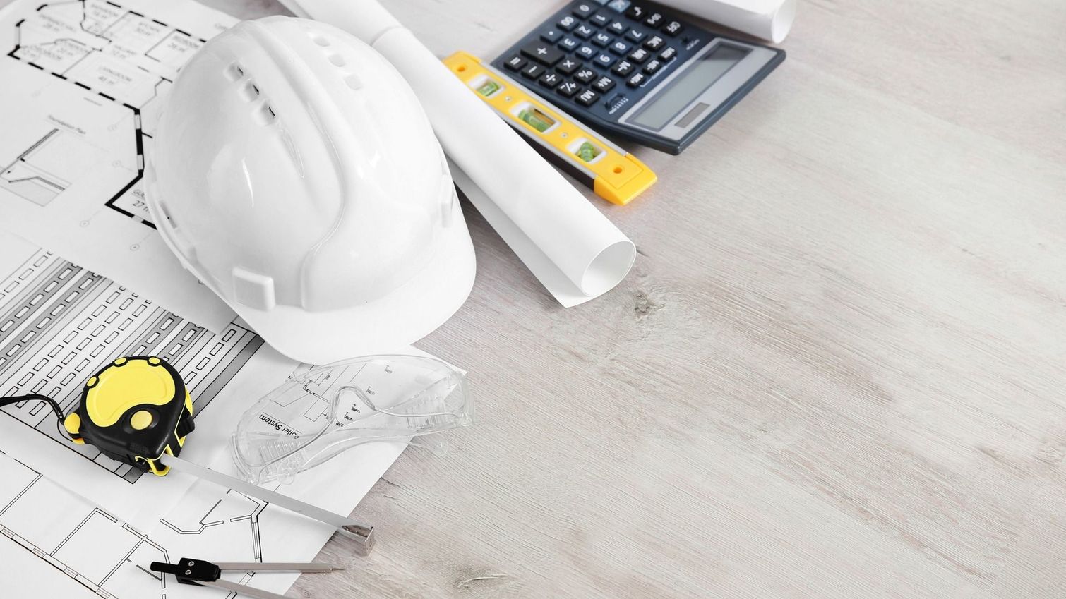 Un elmetto bianco, dei progetti, un metro a nastro, una livella a bolla e una calcolatrice su una superficie di legno chiaro.