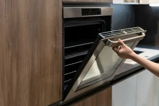 A hand opens the glass door of a modern, stainless steel built-in oven set into dark wood cabinetry.