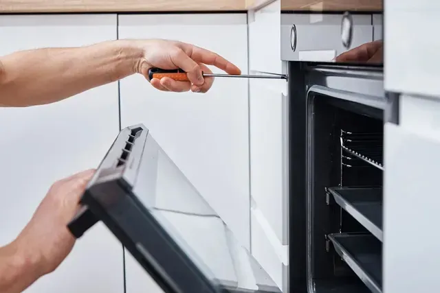 A technician uses a screwdriver to repair the hinge of an open wall-mounted oven in a kitchen.