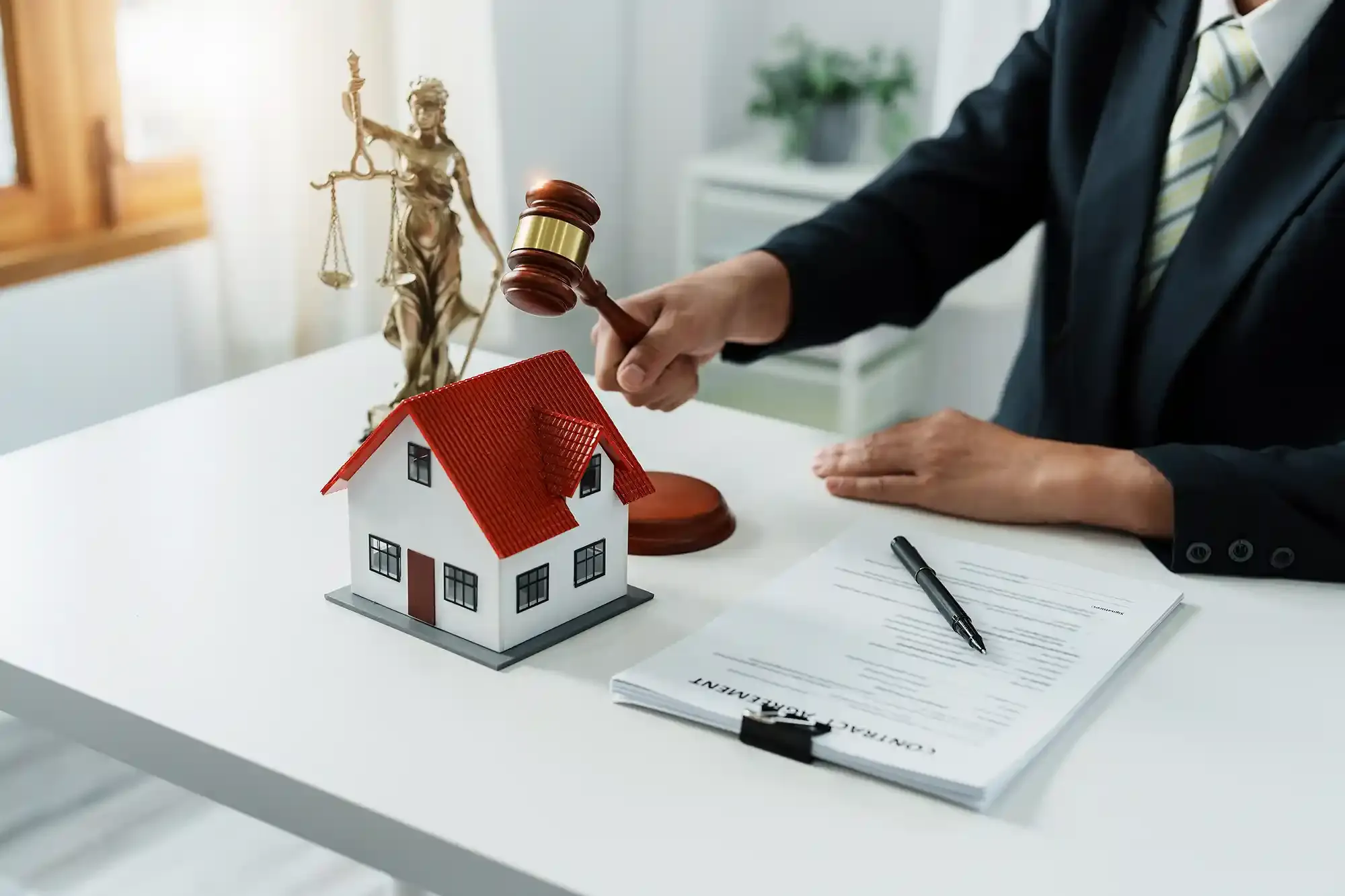 Person in suit using gavel on a model house, beside scales of justice and paperwork.