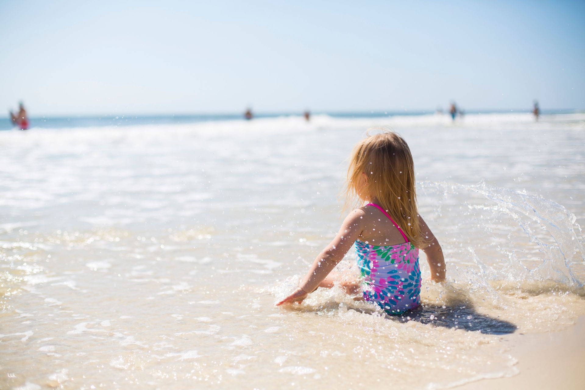 Child sitting in shallow ocean water on a sunny beach, wearing a colorful swimsuit