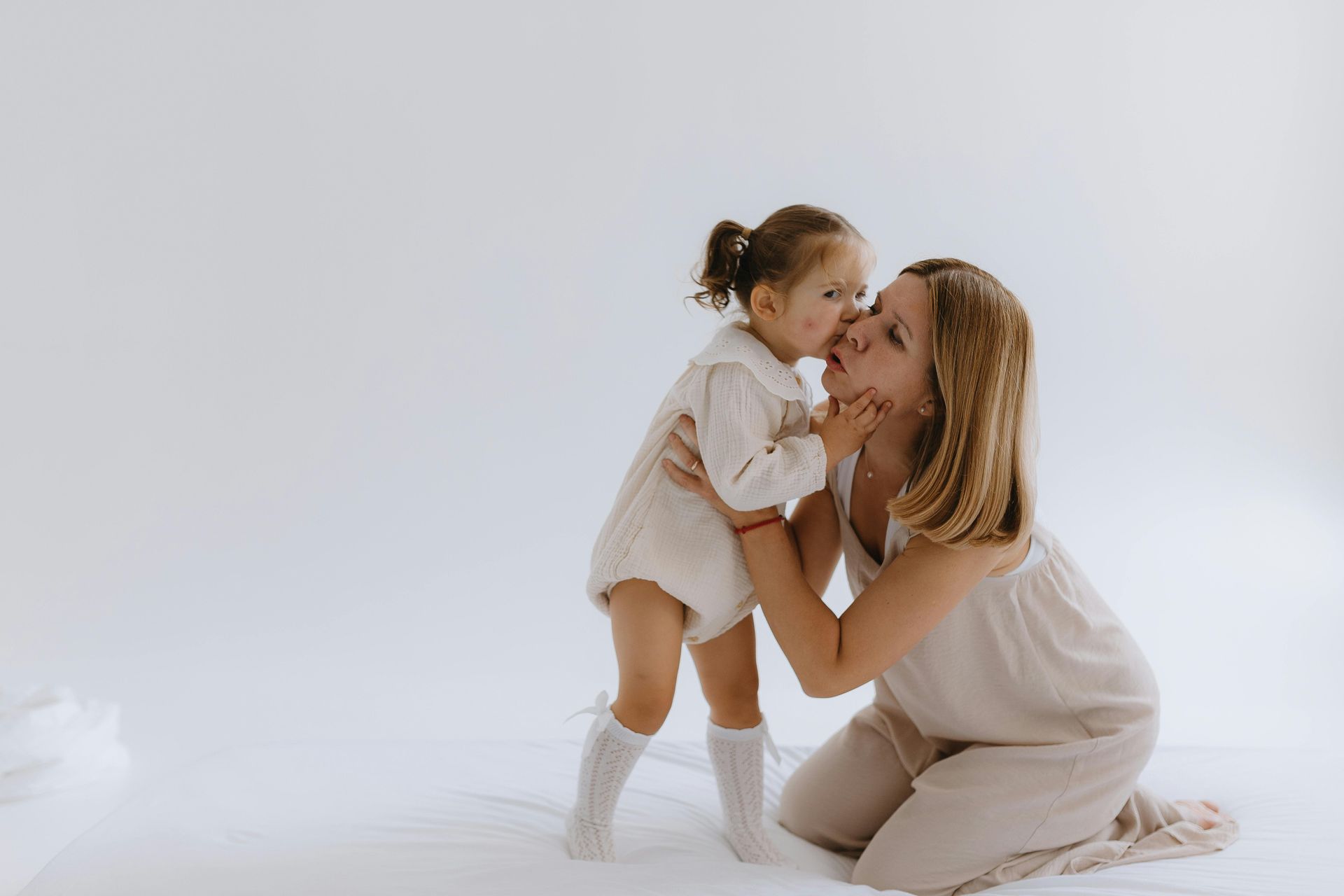 Girl kisses a woman on the cheek. Both are in neutral-colored clothing against a white background.