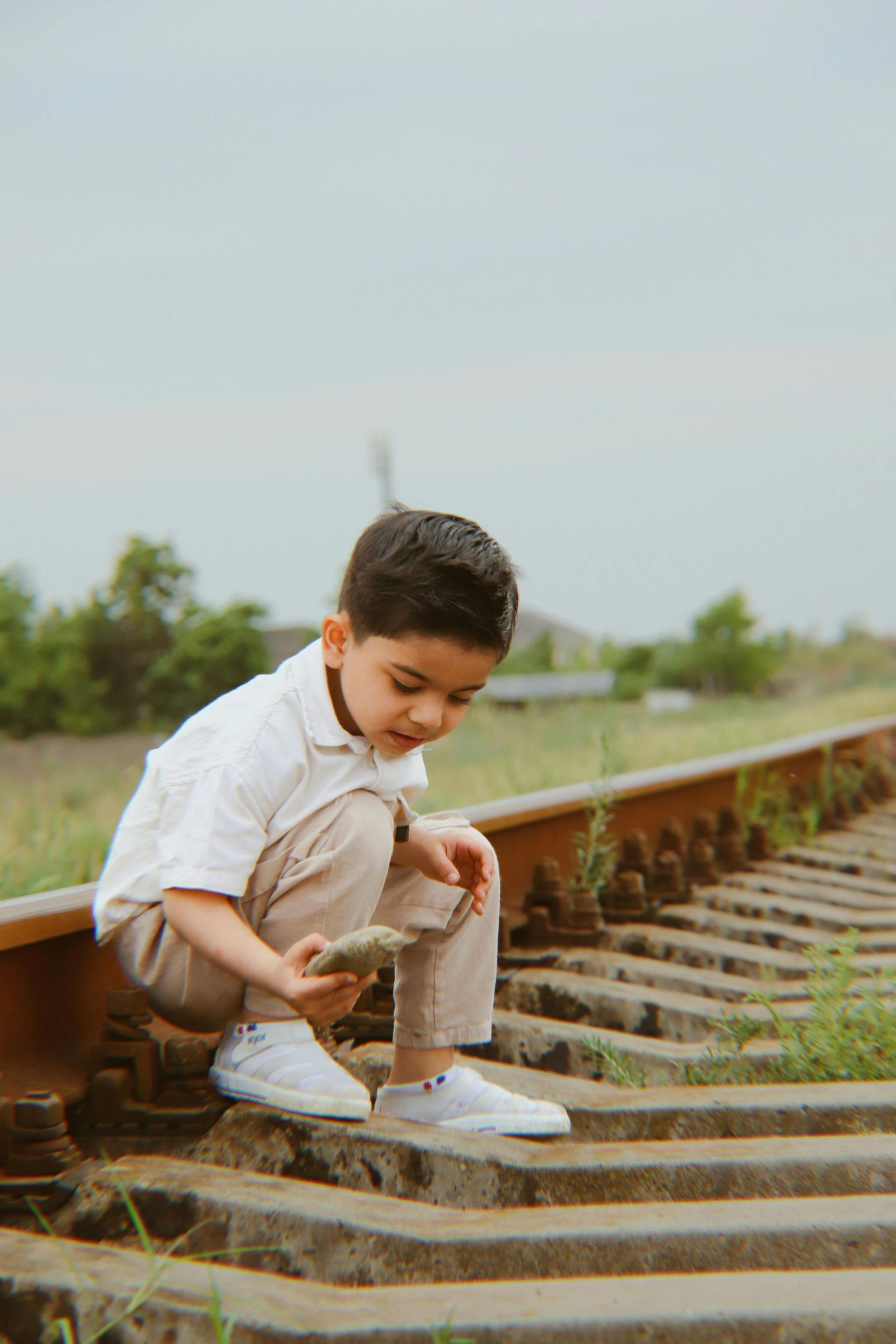 Boy squatting on railroad tracks, examining a rock; outdoors, overcast sky.