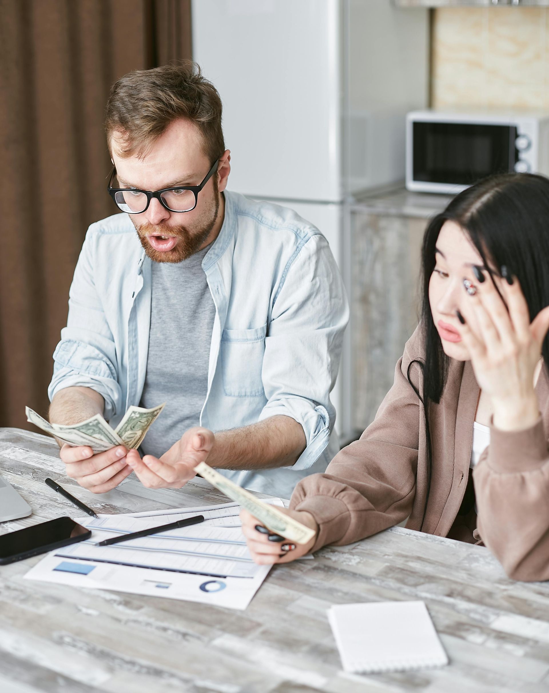 Two people at a table counting cash and reviewing bills, with one looking stressed in a bright office kitchen.