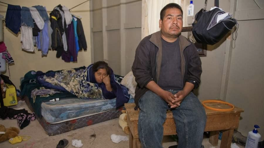 A man is sitting in a messy room with a woman sleeping on a bed.