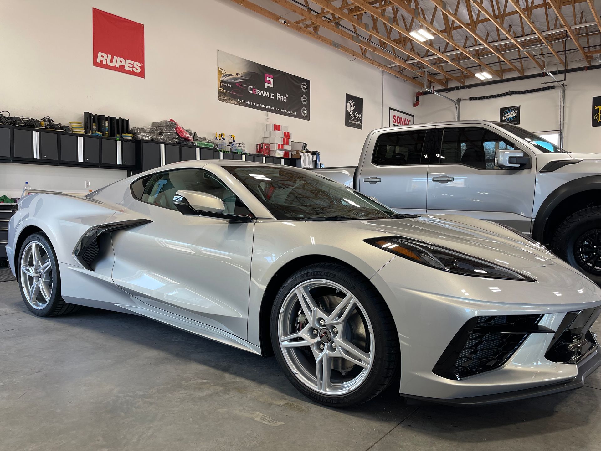 A silver corvette is parked in a garage next to a truck.