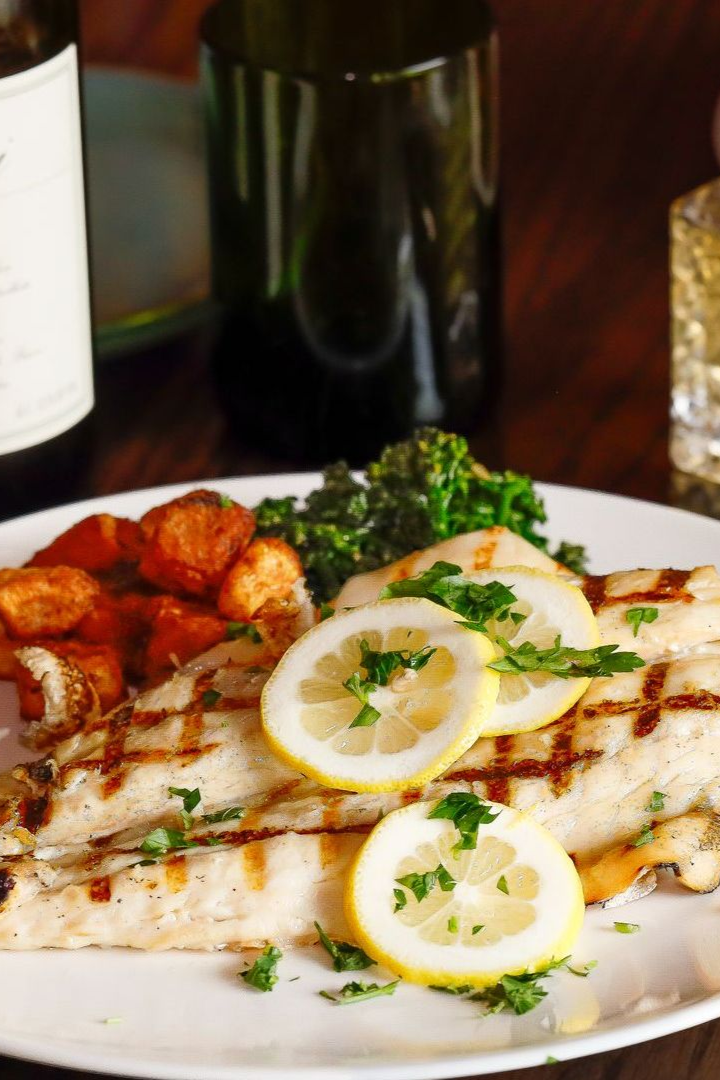 Grilled fish with lemon slices, roasted sweet potatoes, and broccoli rabe on a white plate; wine bottles in background.