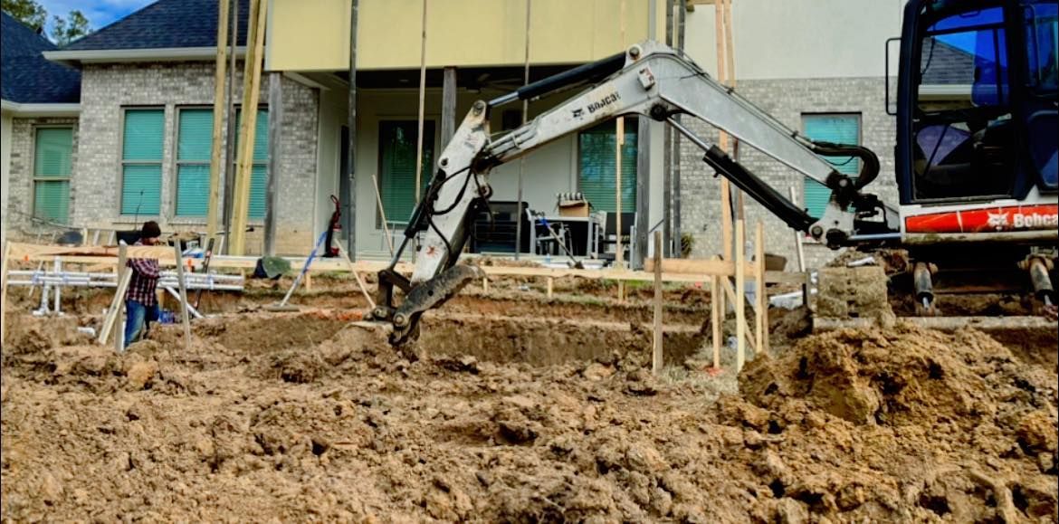 An excavator is digging a hole in the dirt in front of a house under construction.
