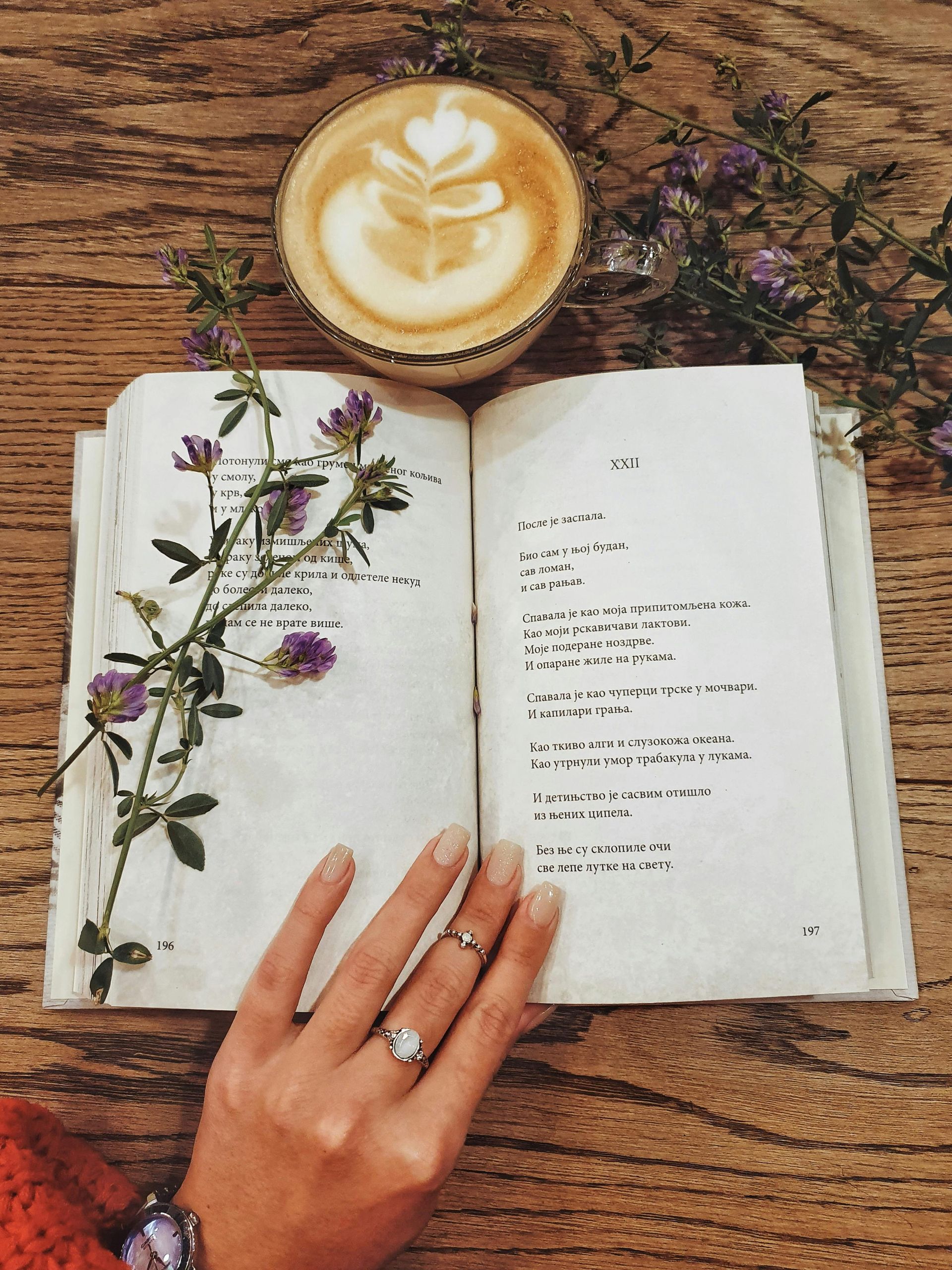 A woman is reading a book next to a cup of coffee and flowers.