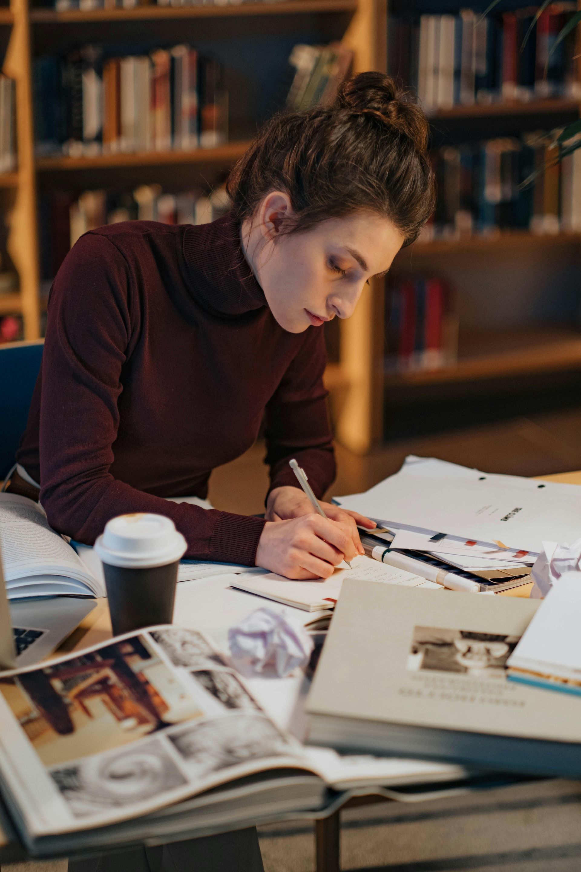 A woman is sitting at a table in a library writing in a notebook.