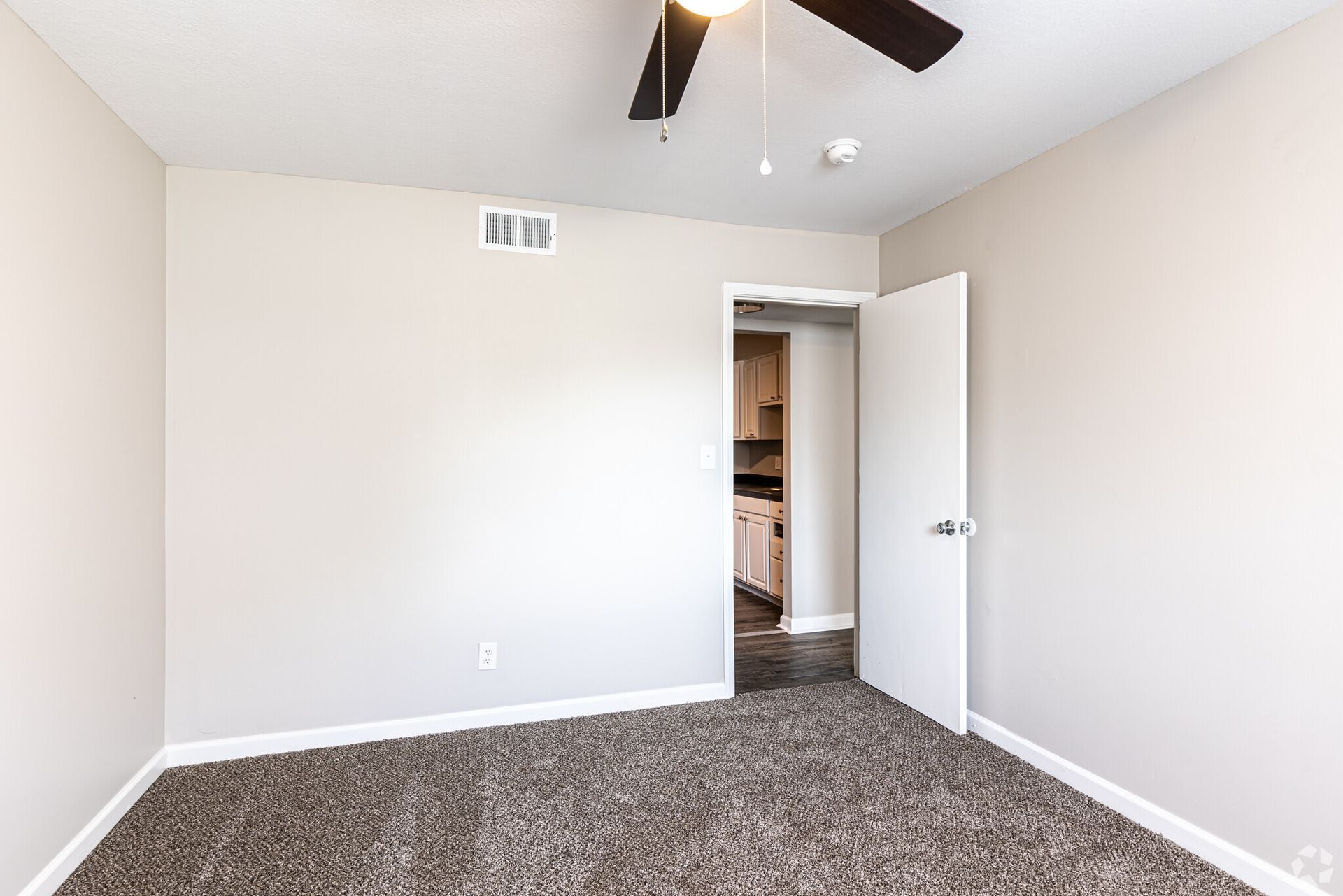 An empty bedroom with a ceiling fan and a door leading to a kitchen.