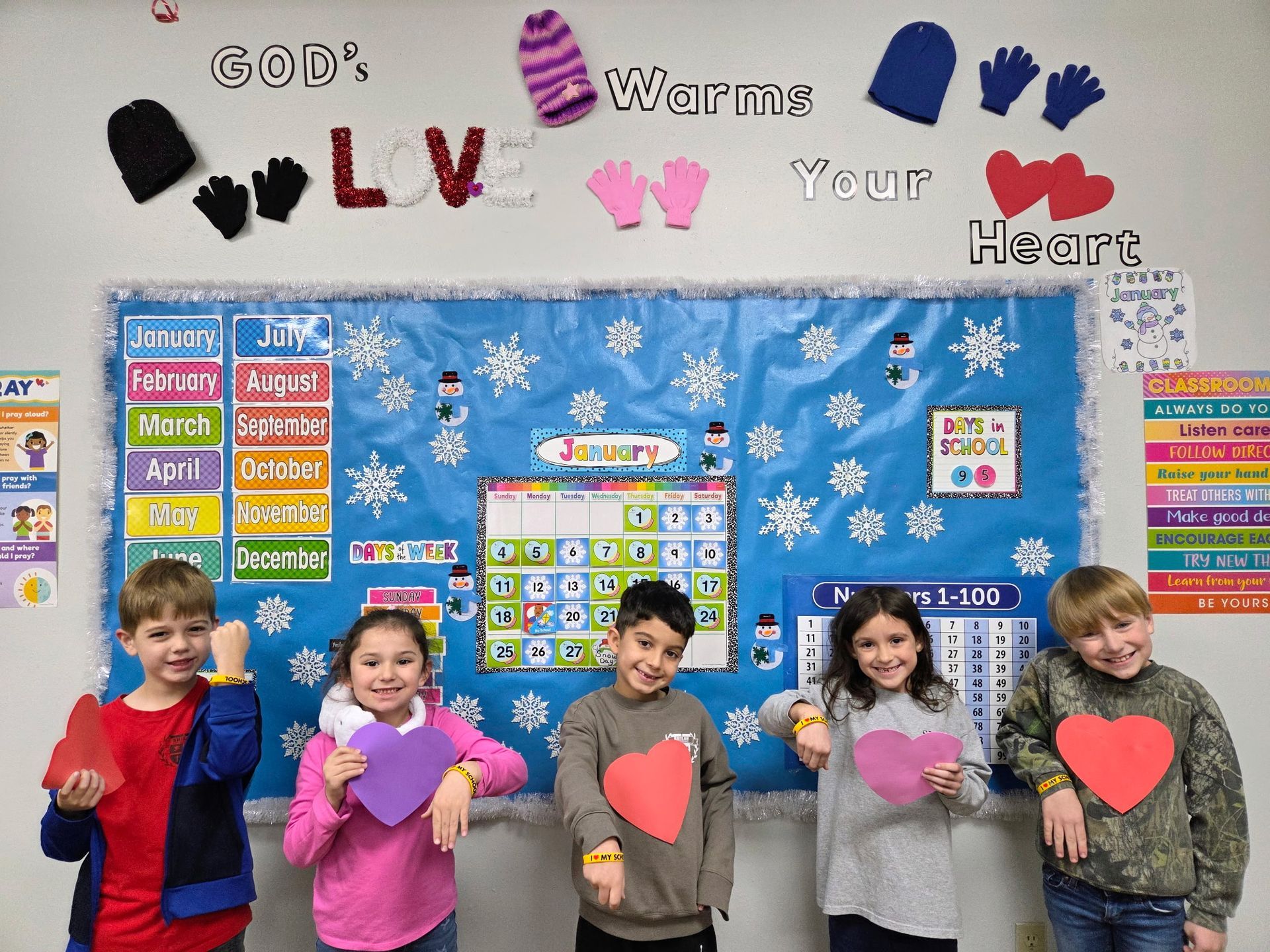 Children holding heart cutouts in front of a classroom bulletin board decorated with winter and religious themes.