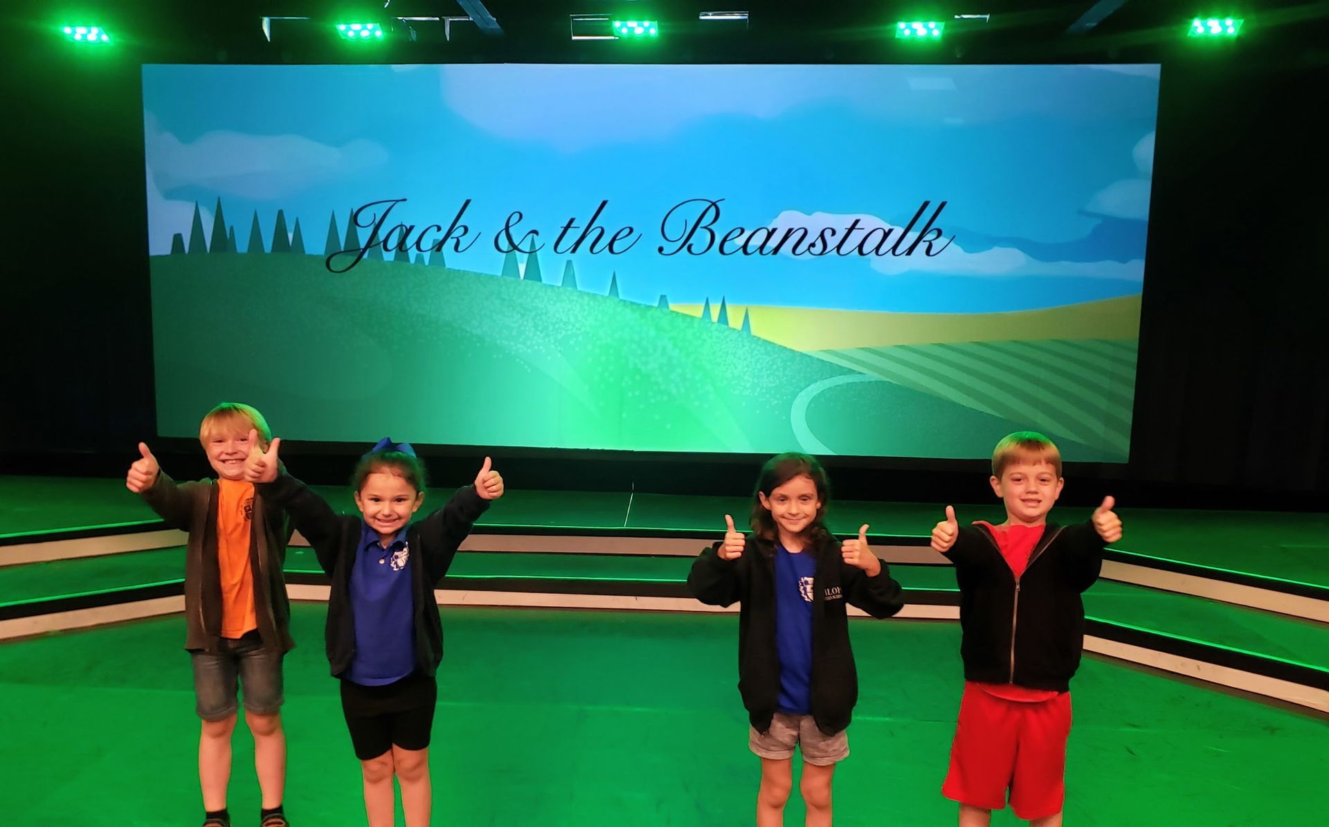 Four children on stage with thumbs up, in front of a backdrop of