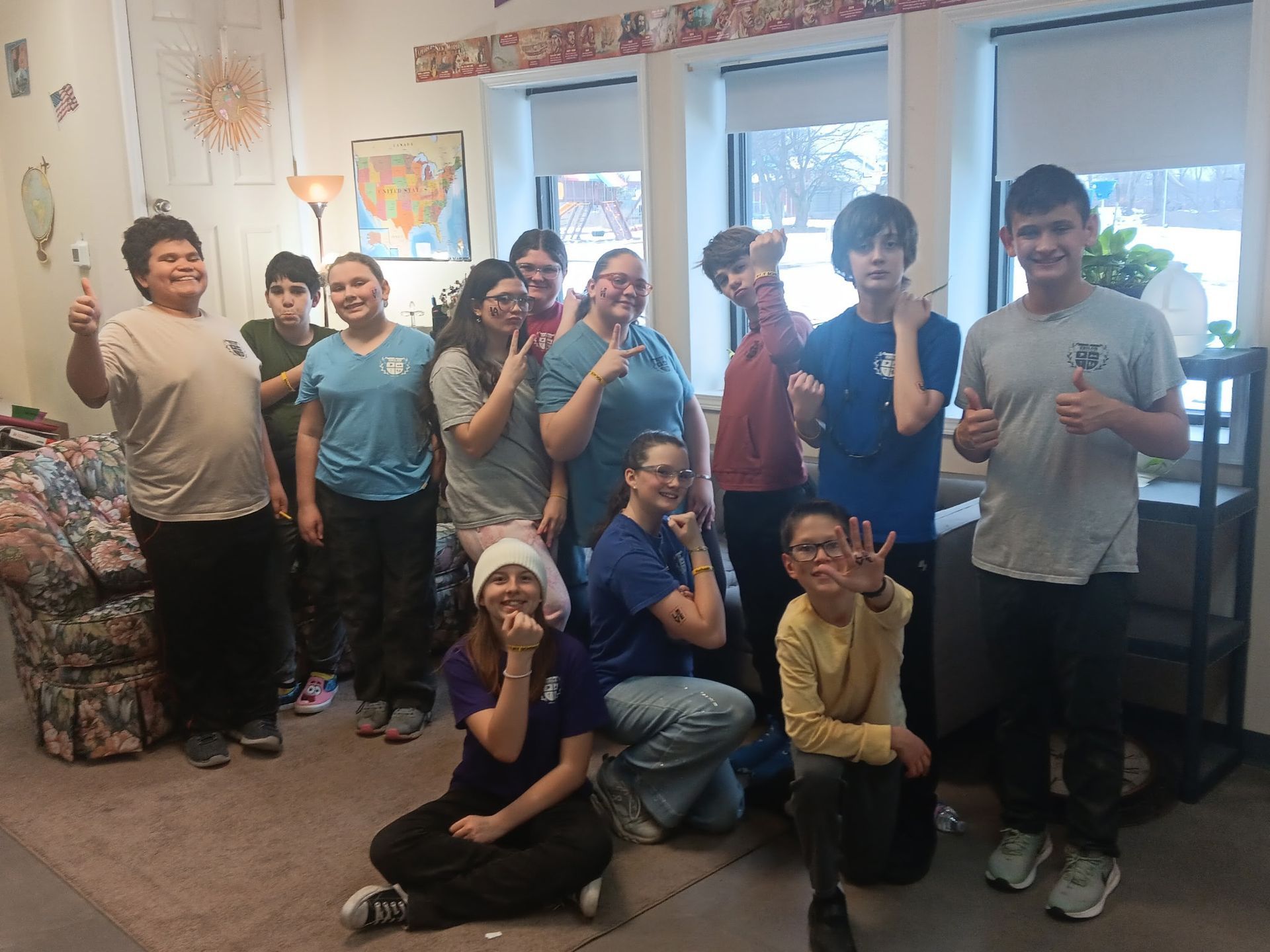 Group of people smiling, making hand gestures in a brightly lit room with a window; some wearing matching t-shirts.