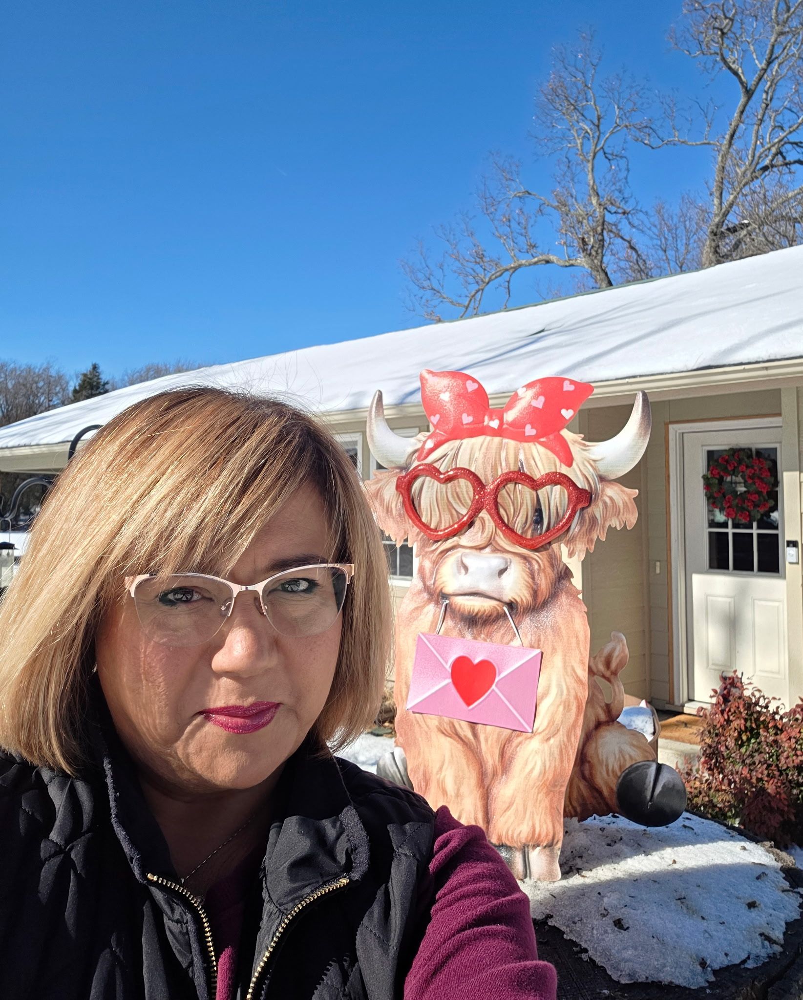 Woman with glasses taking a selfie next to a Valentine's Day decorated Highland cow statue outside a snowy house.