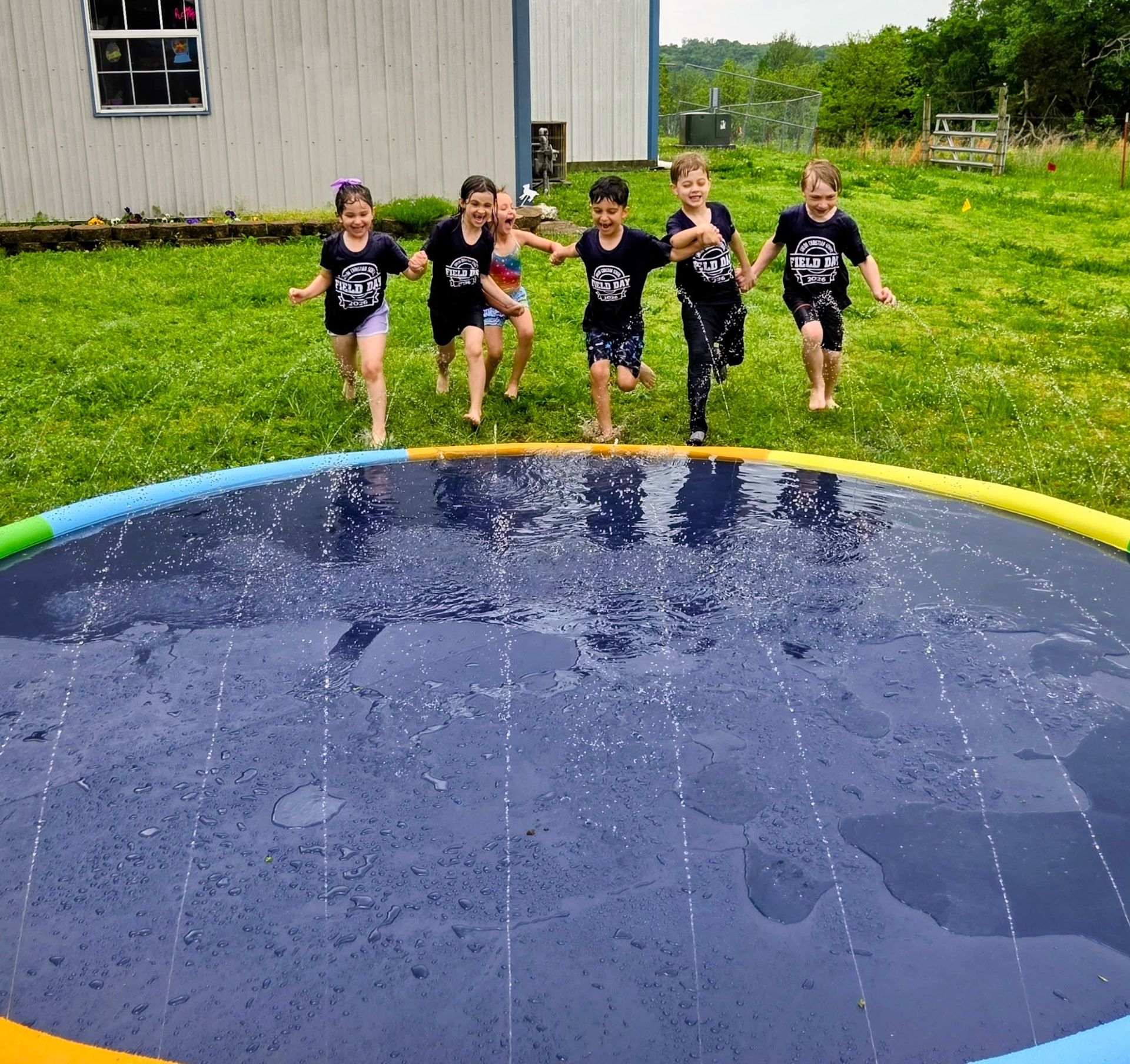 Five children in black shirts jumping into a blue splash pool on a grassy day camp field