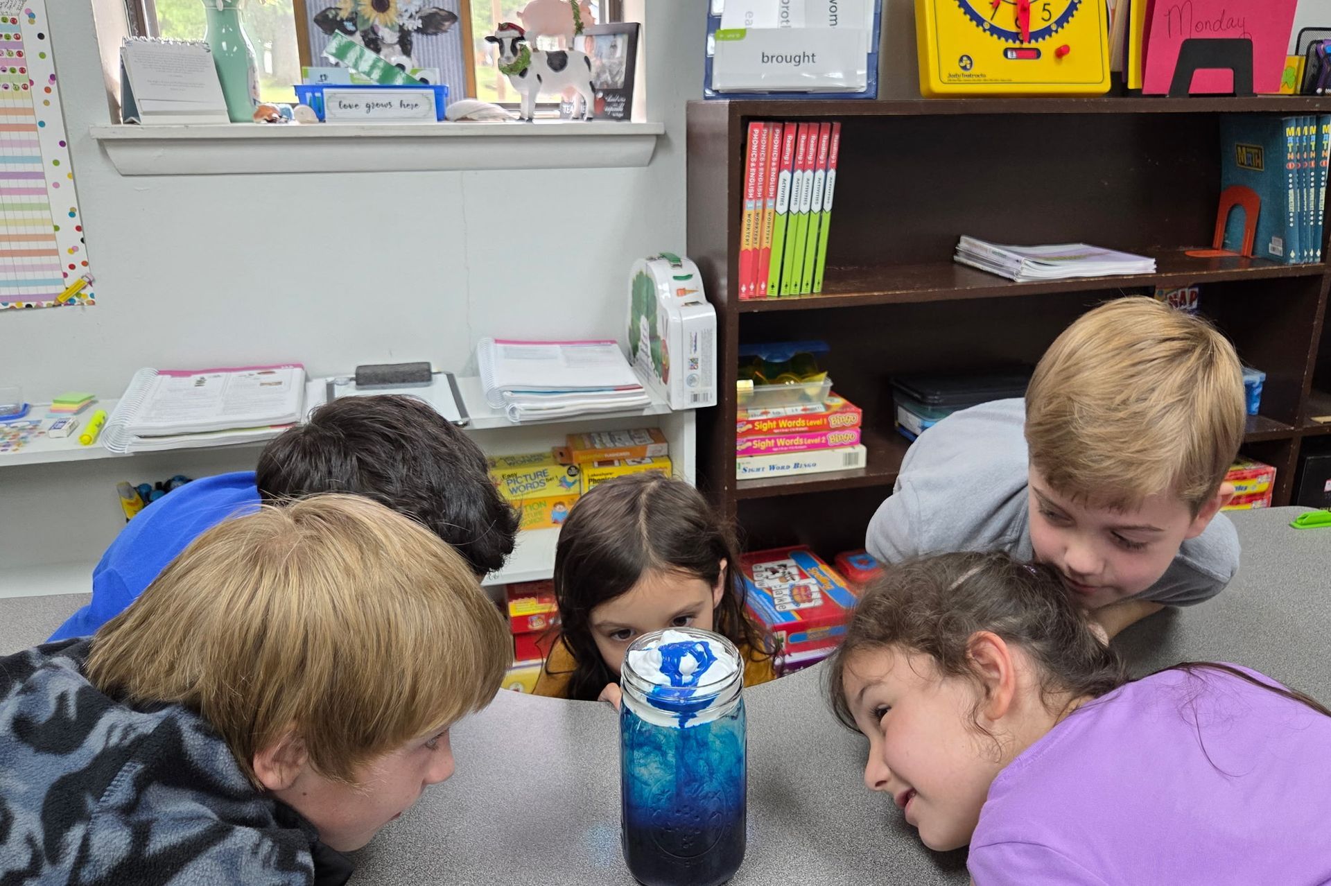 Children gathered around a table, peering at a blue water bottle in a classroom.