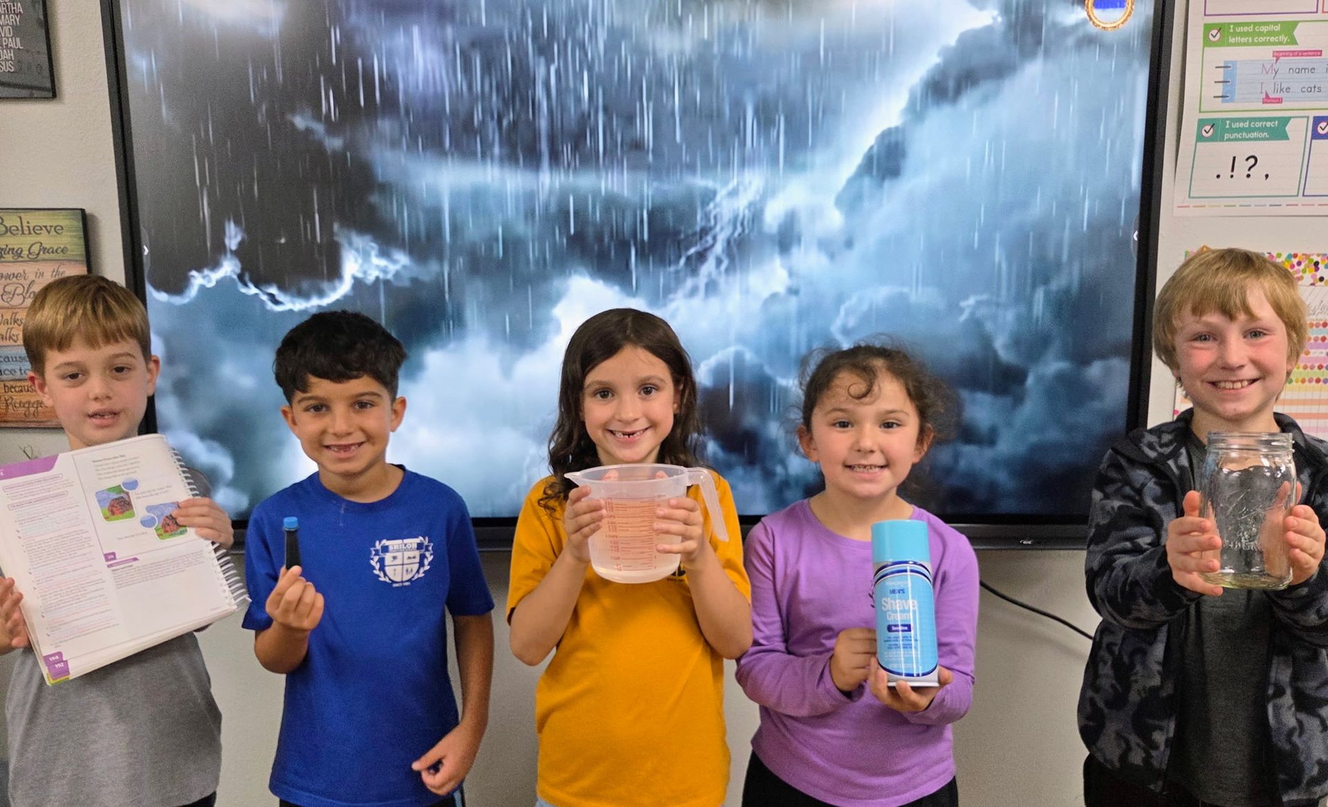 Five children in a classroom hold up artwork and bottles in front of a large screen.