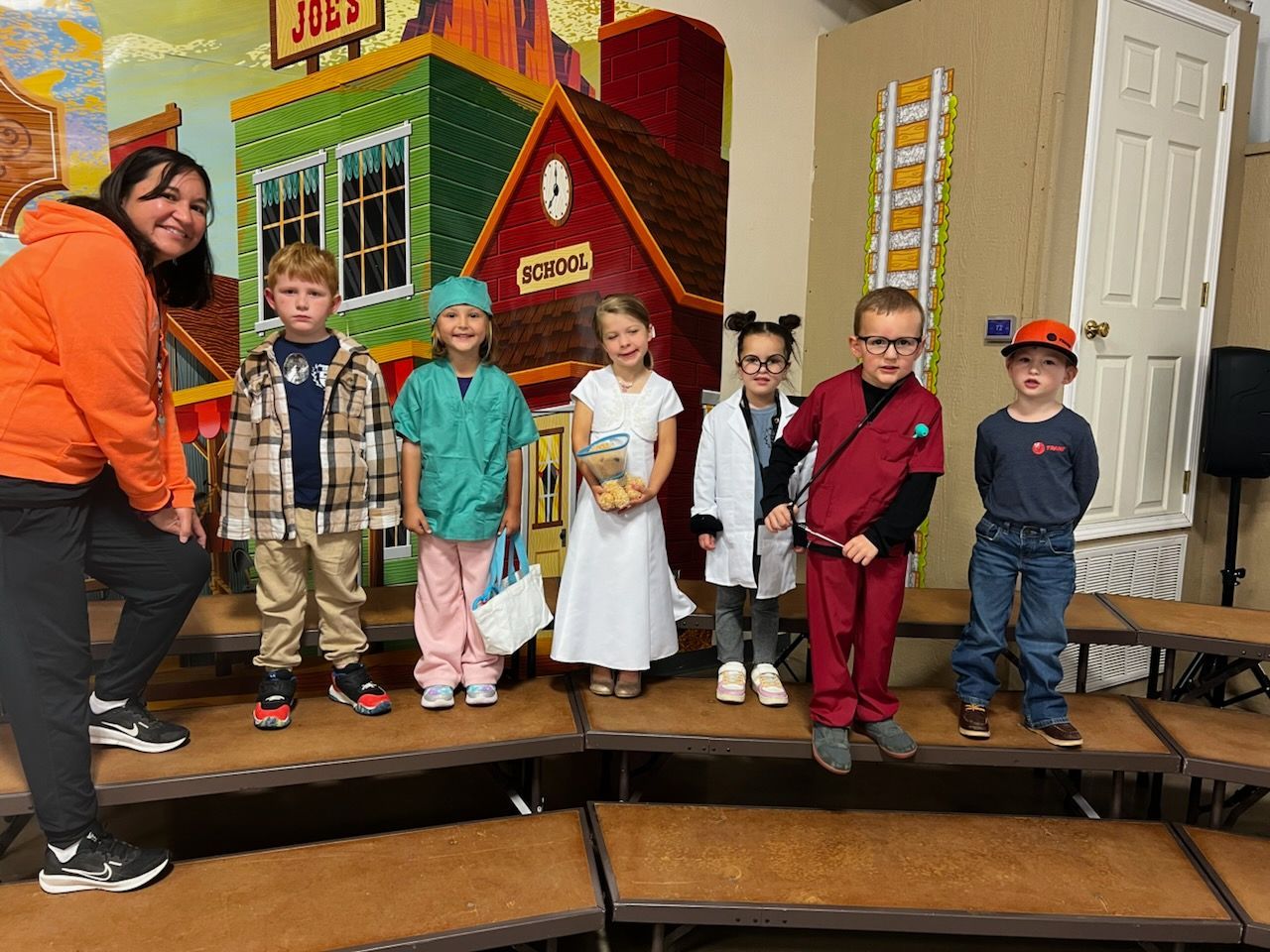 Teacher with children in costumes on stage; cowboy, doctor, bride, scientist, and more.