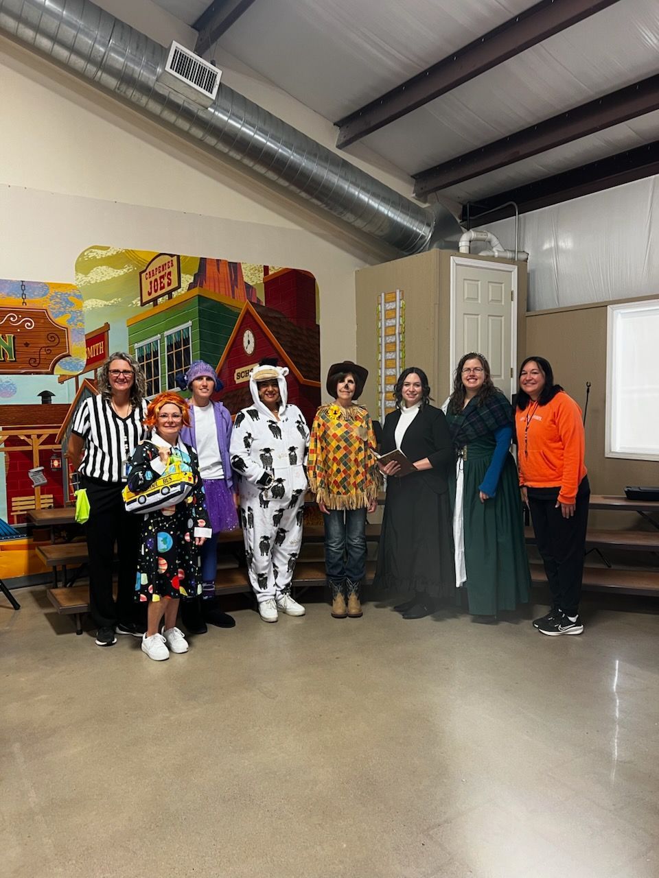 Group of people in costumes posing, staged indoors. Colorful backdrop with building mural.