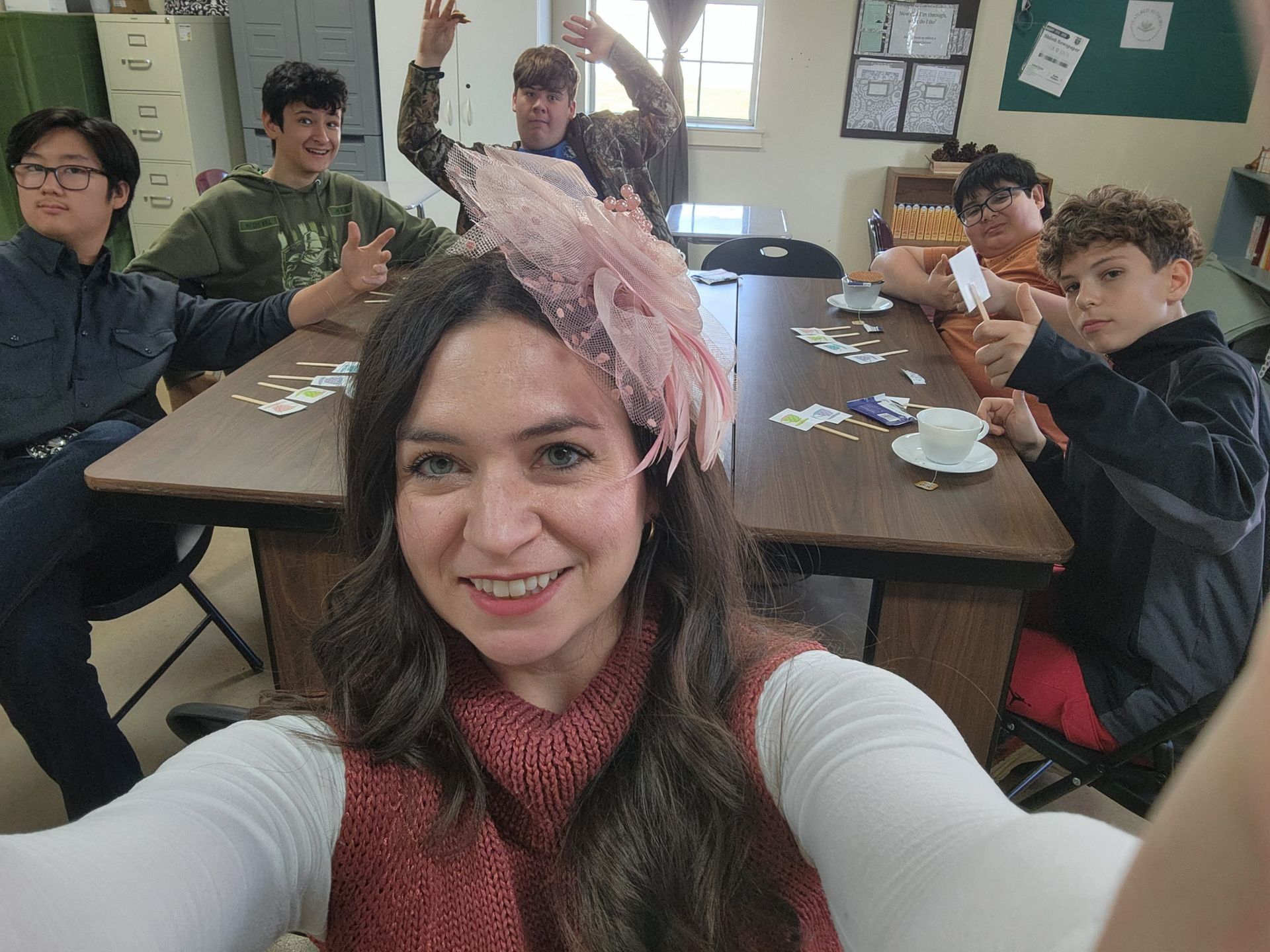 Woman in hat smiles, taking a selfie with a group of seated people at a table, gesturing and holding cards. Room setting.