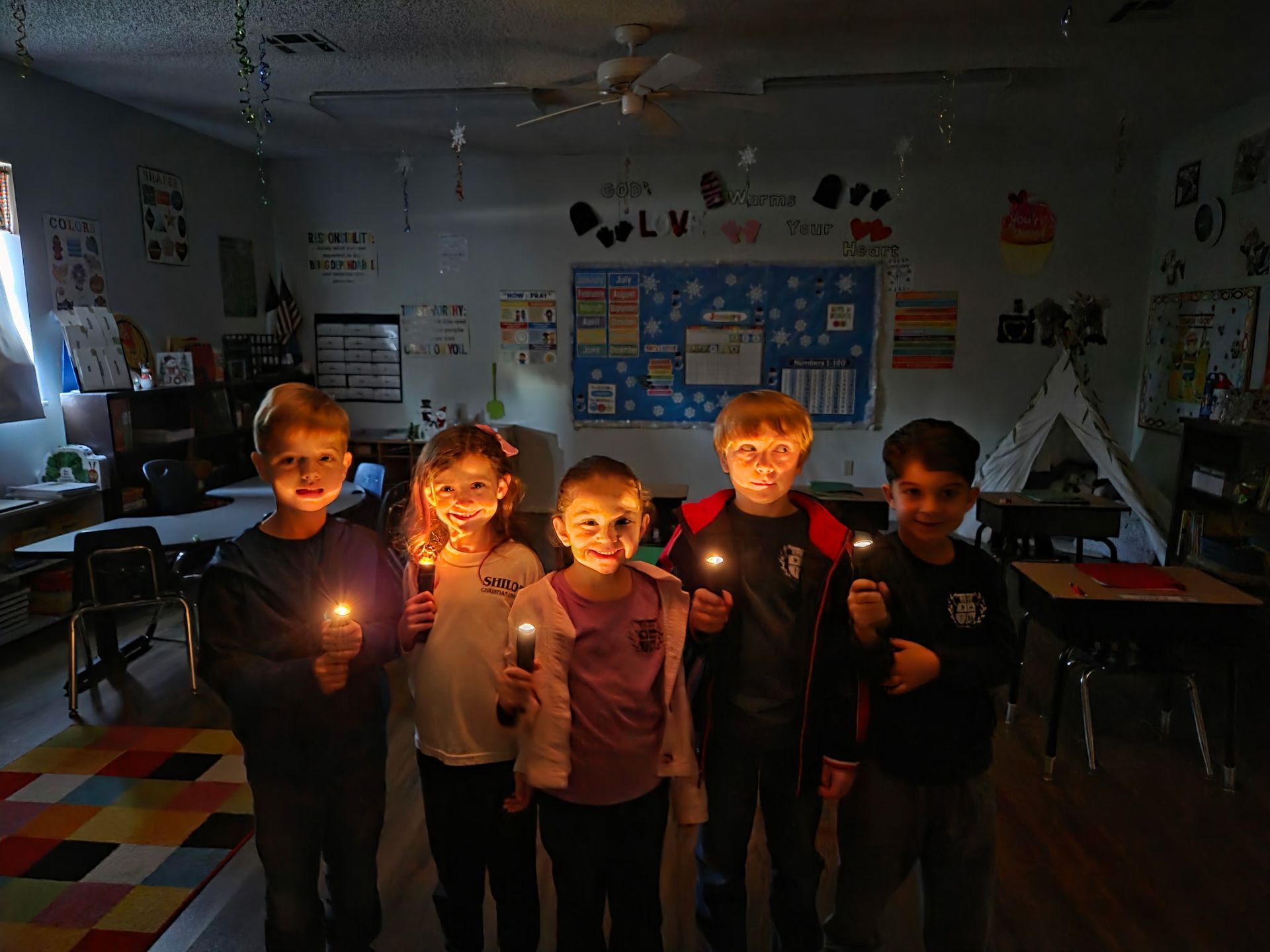 Five children in a classroom hold lit candles in dim lighting.