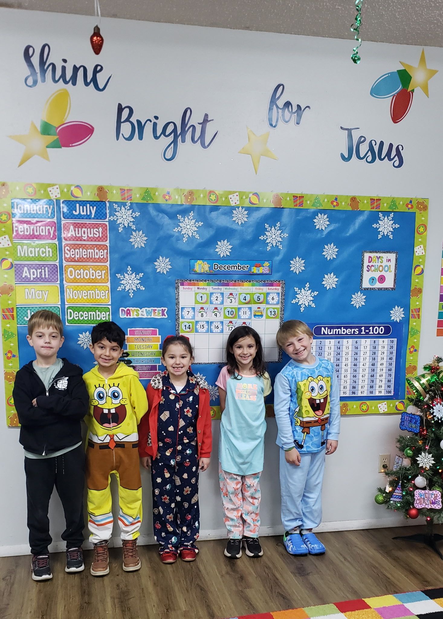 Five children in pajamas stand in front of a classroom display.