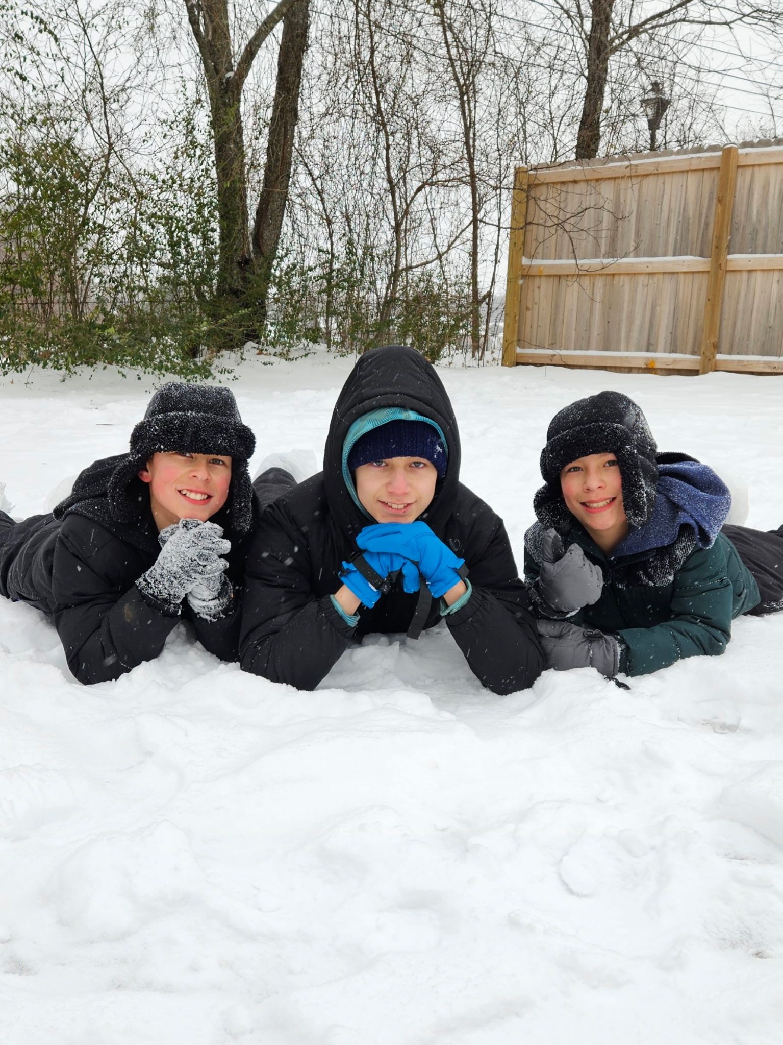 Three people in winter jackets and hats lying in the snow, smiling.