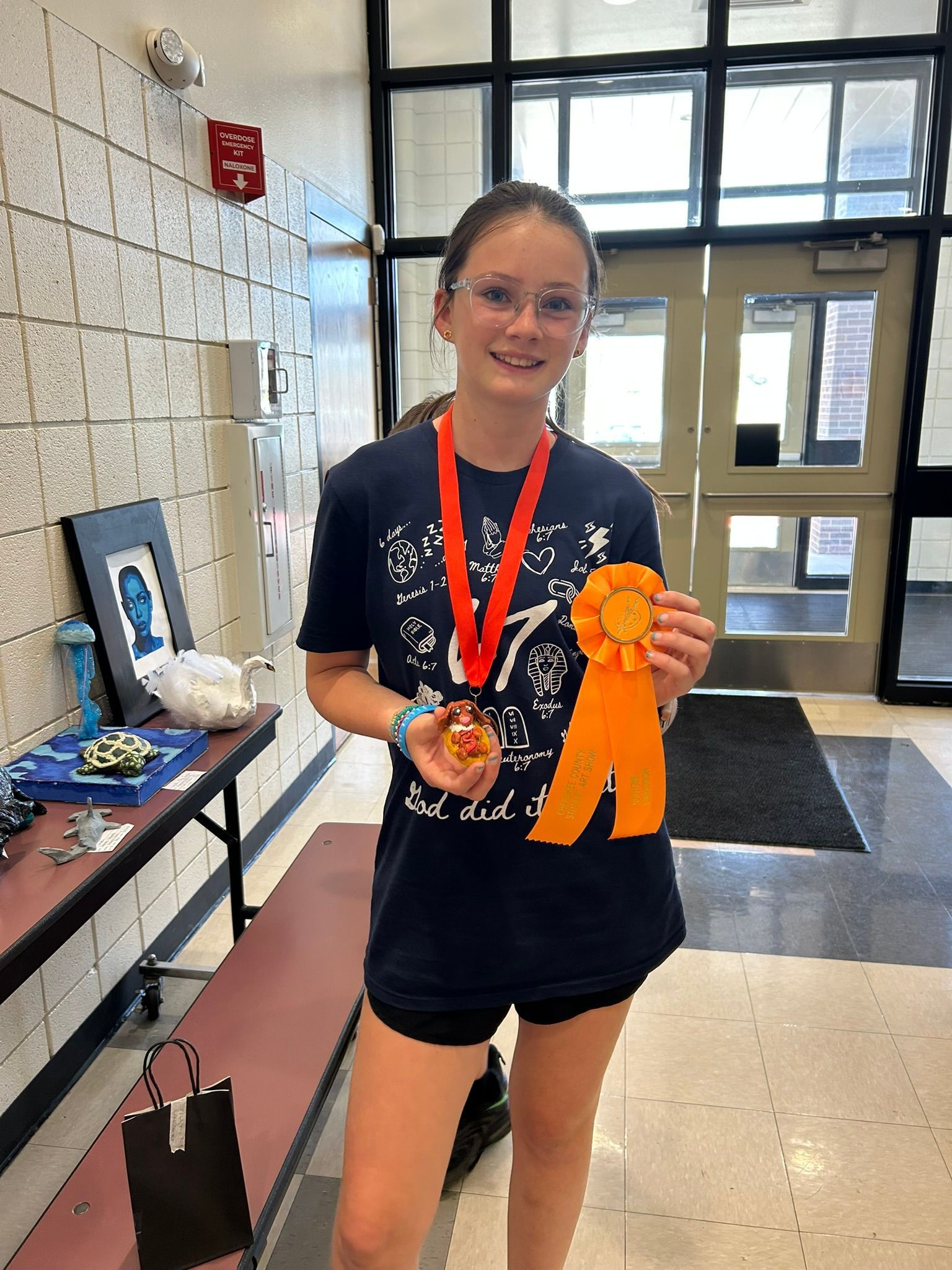 Girl holding an orange ribbon and medal in a school hallway, smiling at the camera.