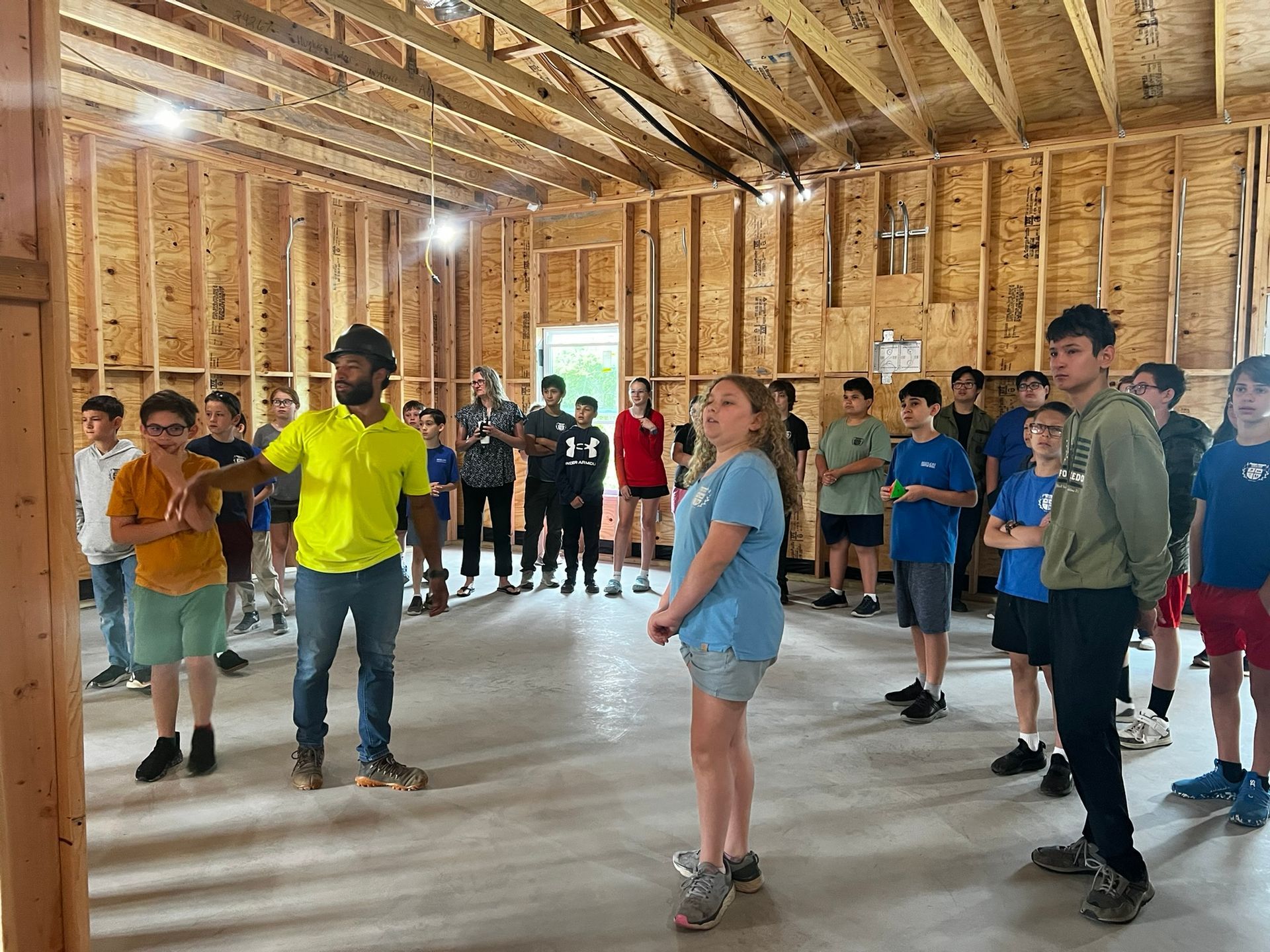 Group standing in an unfinished wooden room, facing an instructor in a bright yellow shirt.