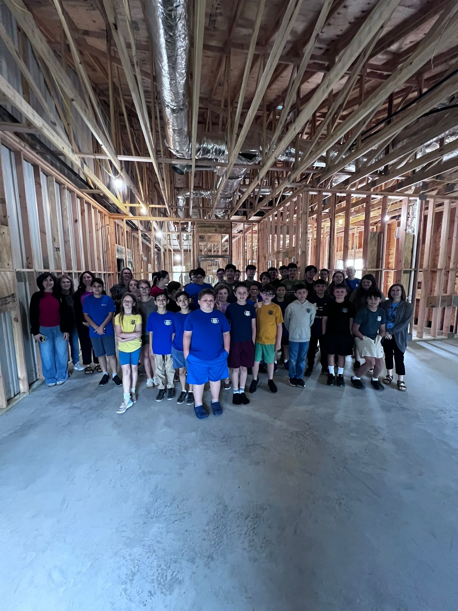 Group of children and adults standing in an unfinished wooden building with exposed beams and concrete floor