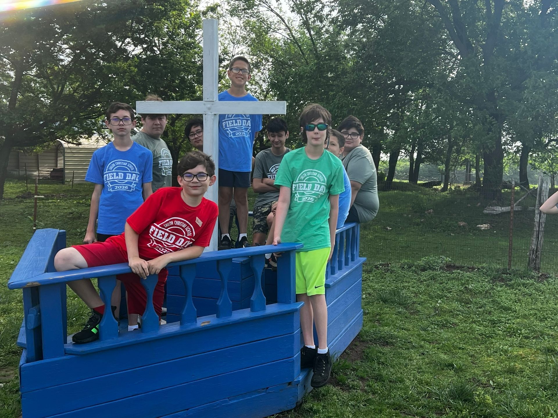 Group of people posing on a blue boat-shaped structure in a grassy park, with trees in the background.
