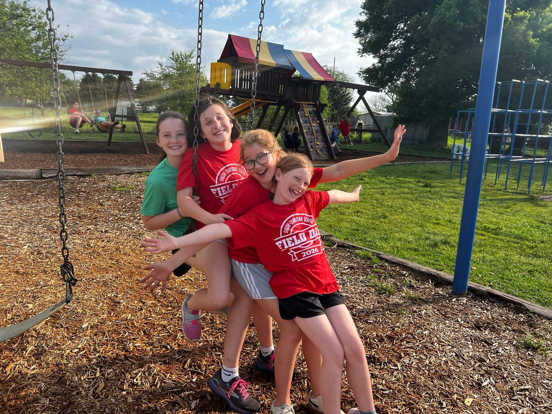 Four children on a playground swing, smiling and leaning together under a sunny sky.