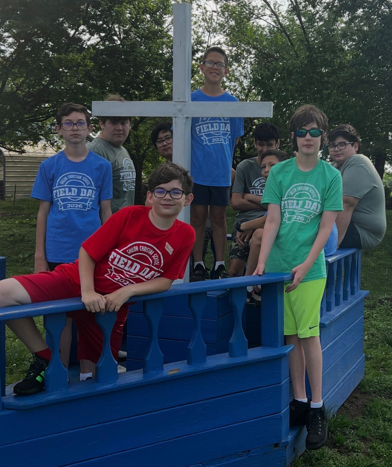 Five teens posing on a blue bridge, wearing bright shirts outdoors among trees.